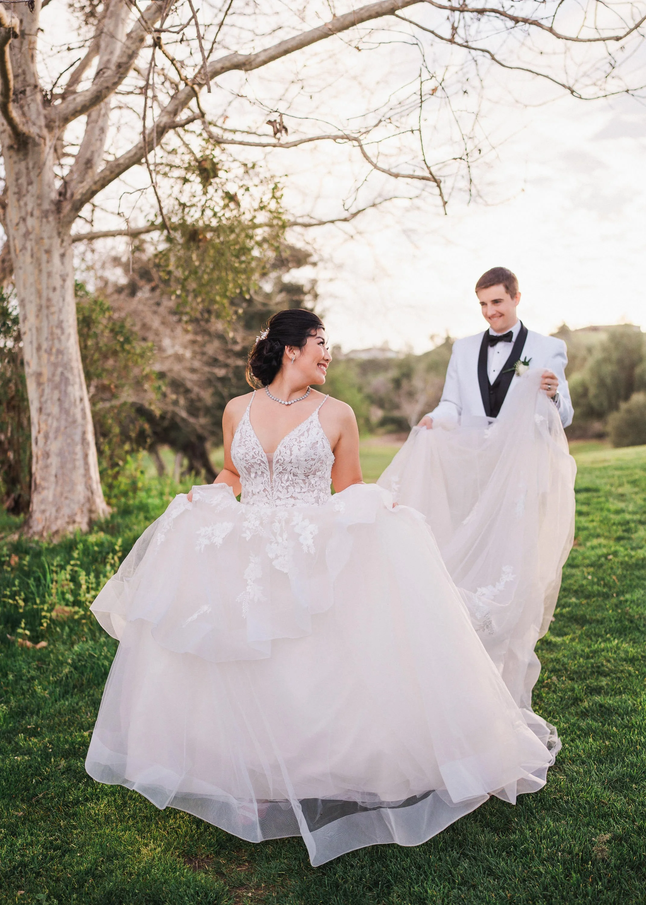 A bride and groom on their wedding day outdoors, with the groom holding up the bride’s wedding dress as they smile at each other.