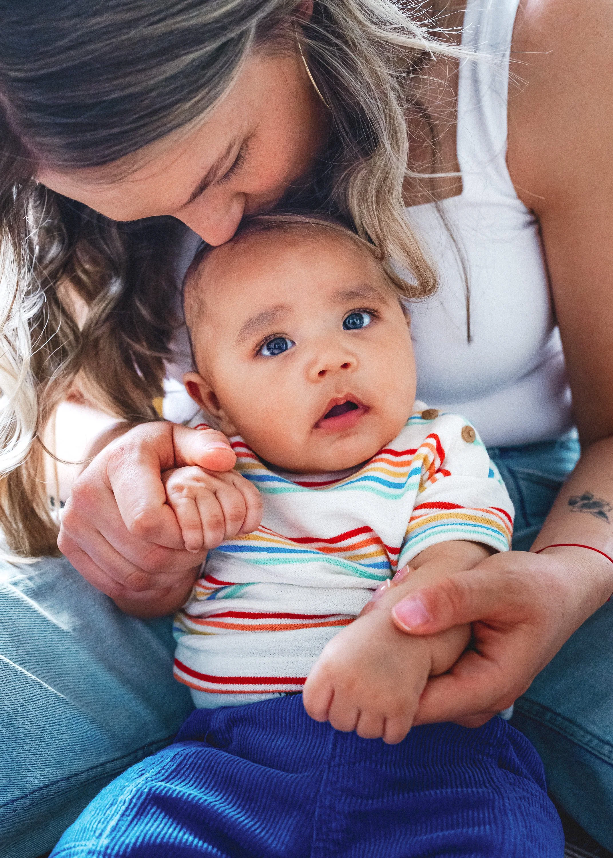 A woman with wavy blonde hair leaning over an infant, gently holding the baby's hand, with the woman kissing the baby's head. The baby has blue eyes, light brown hair, and is wearing a striped shirt and blue pants.