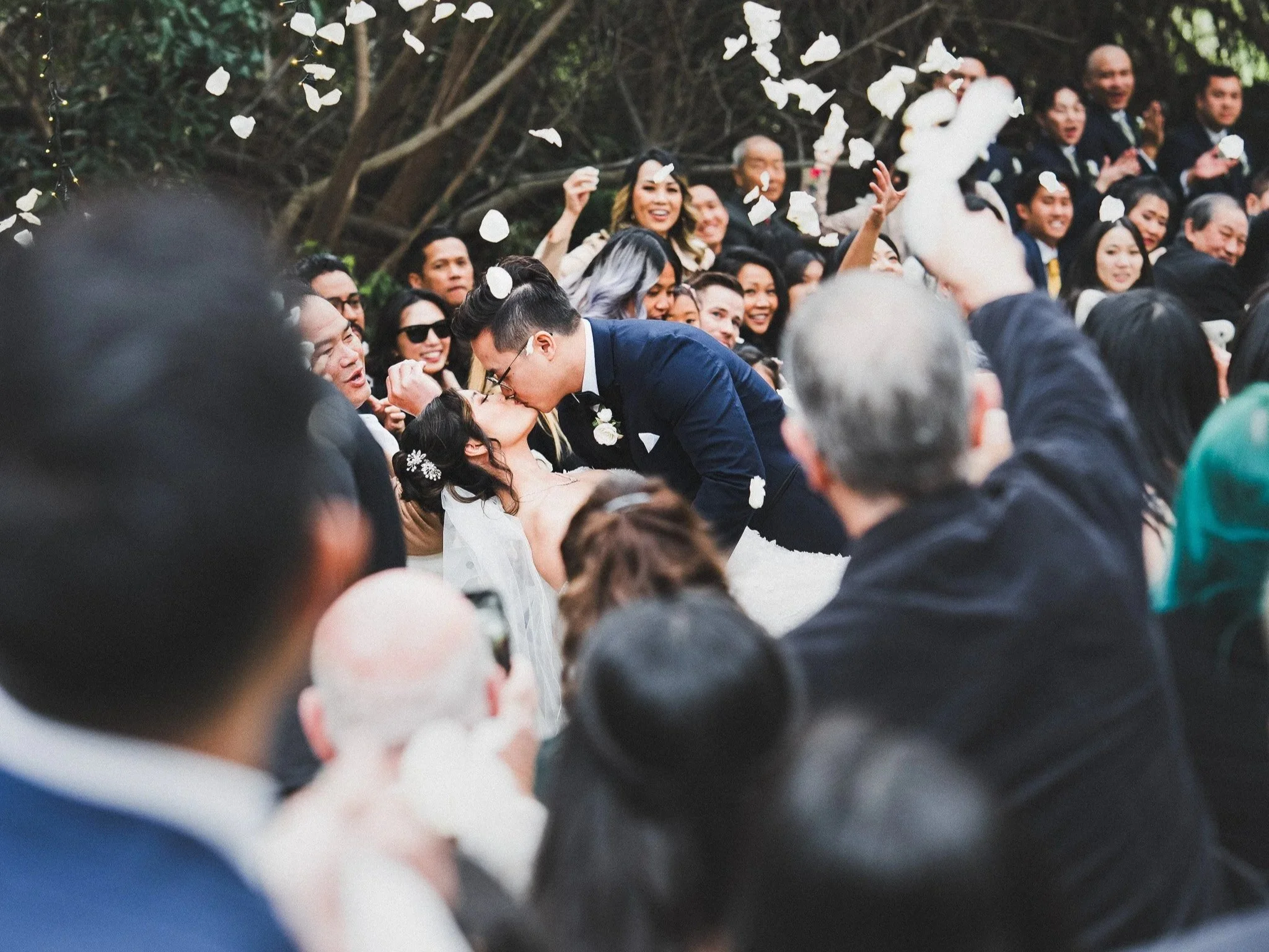 A wedding couple kissing surrounded by wedding guests throwing flower petals in the air.