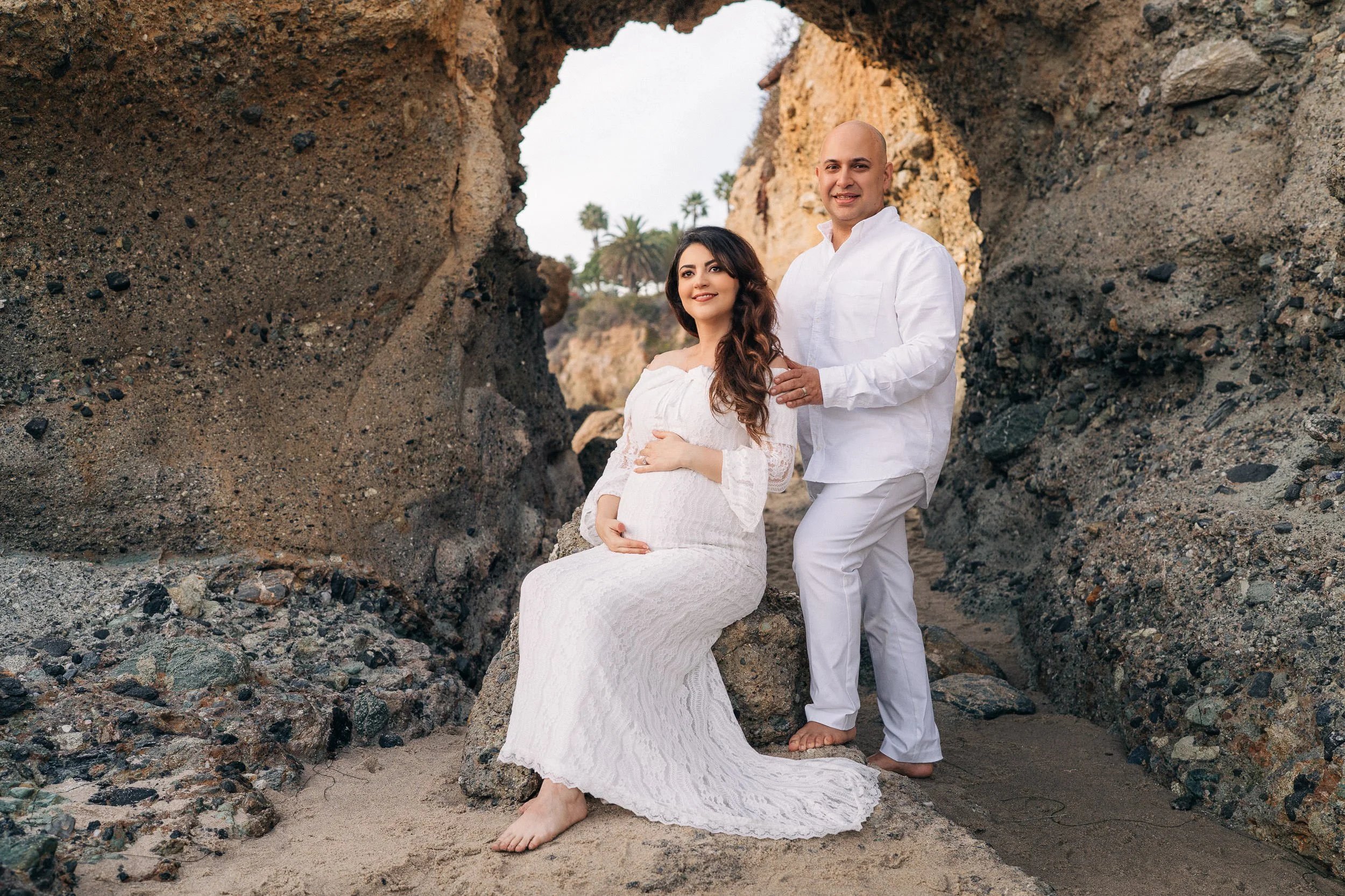 A pregnant woman and a man are standing on a beach near a rocky arch. The woman is seated on a rock, wearing a white lace dress, and has her hand on her belly. The man is standing behind her, also dressed in white, with one hand on her shoulder. Both are smiling and looking at the camera, with palm trees visible in the background.
