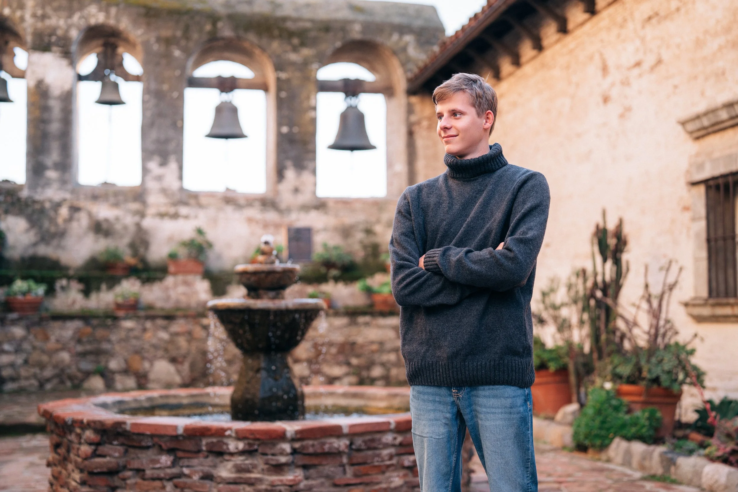 A young man standing outdoors with his arms crossed, wearing a dark gray turtleneck sweater and jeans, in front of an old stone fountain and a weathered building with large windows and hanging bells.
