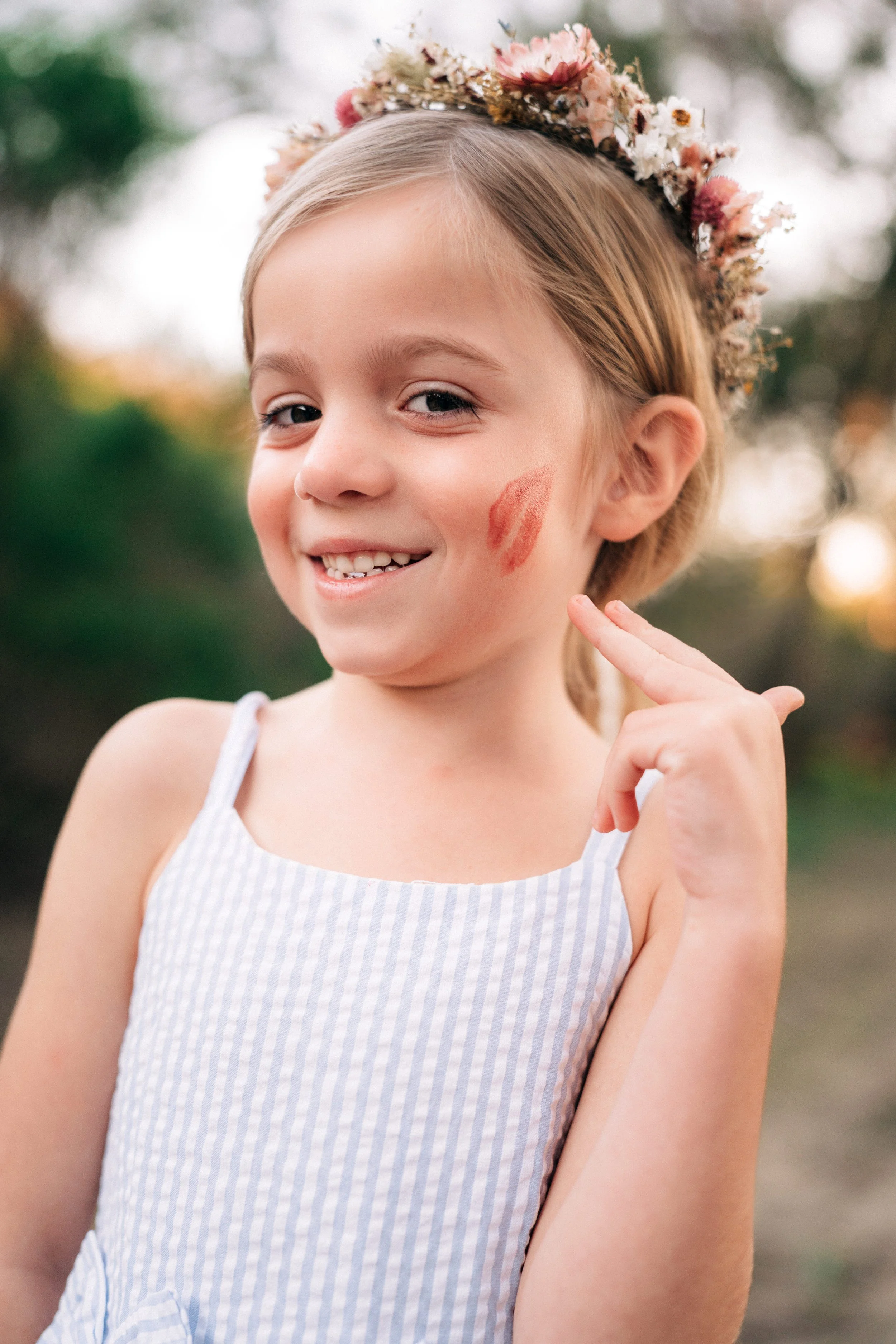 A young girl with a flower crown on her head, smiling and showing a lipstick kiss mark on her cheek, outdoors with blurred greenery in the background.