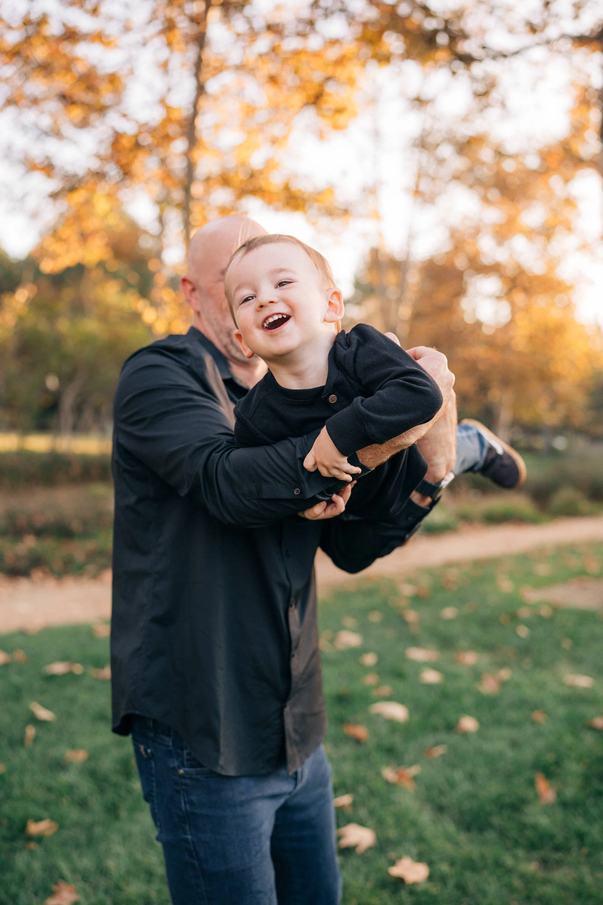 A man holding a laughing young boy outdoors during fall, with trees and fallen leaves in the background.