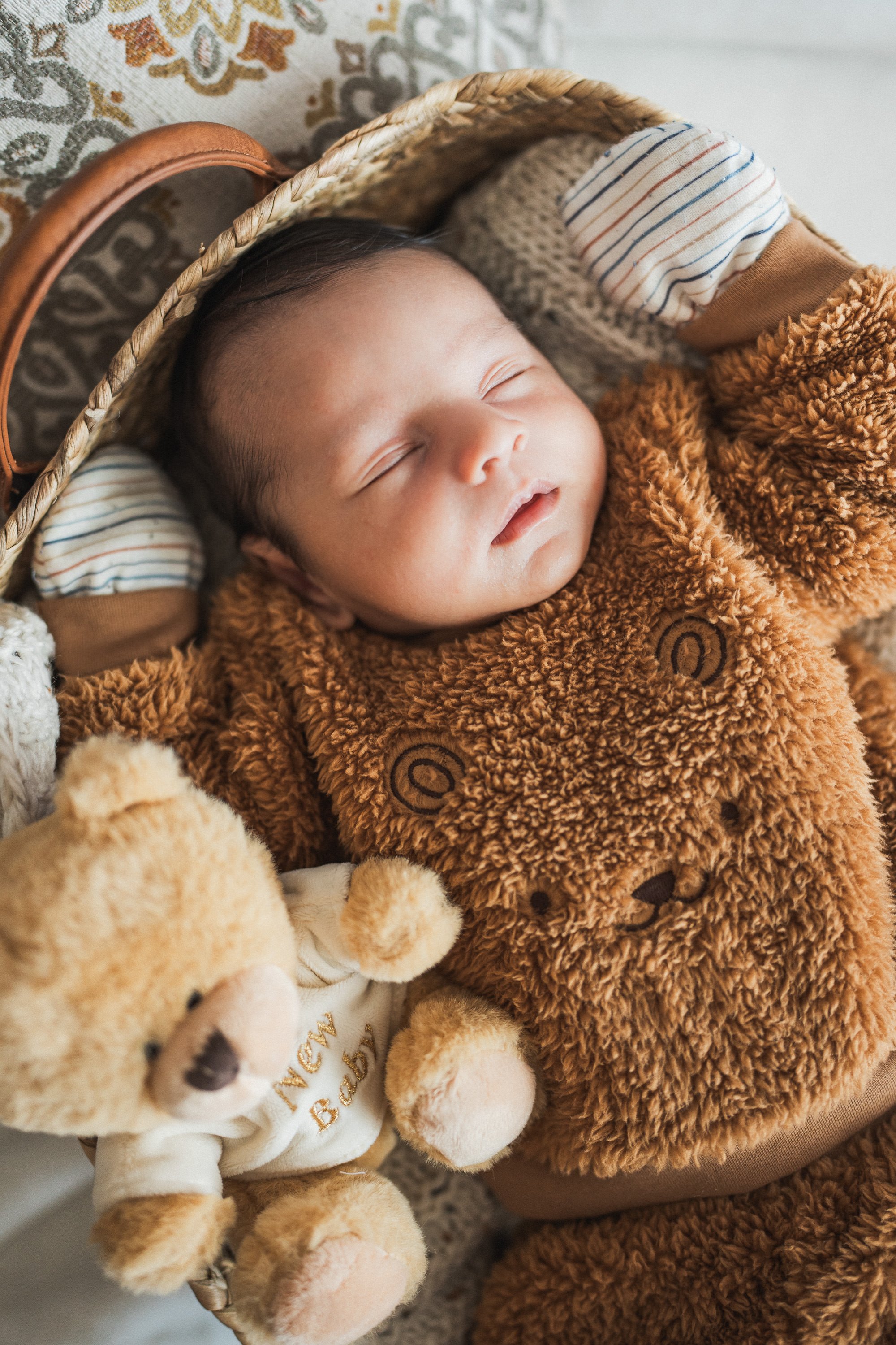 A sleeping baby in a cozy brown teddy bear outfit, lying in a basket, holding a plush teddy bear toy.