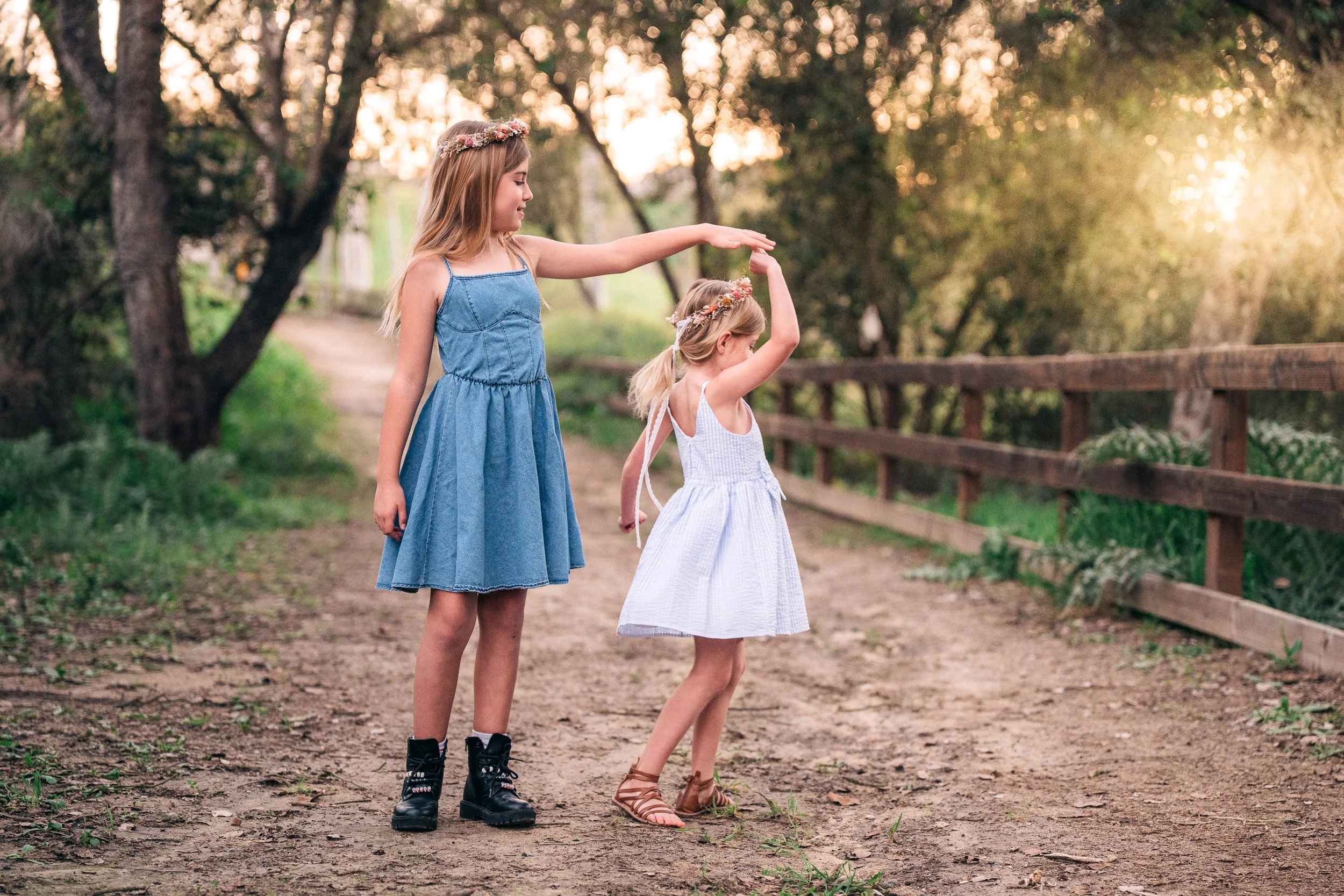 Two young girls in dresses, a taller girl guiding a smaller girl to dance on a dirt path in a wooded area during sunset.