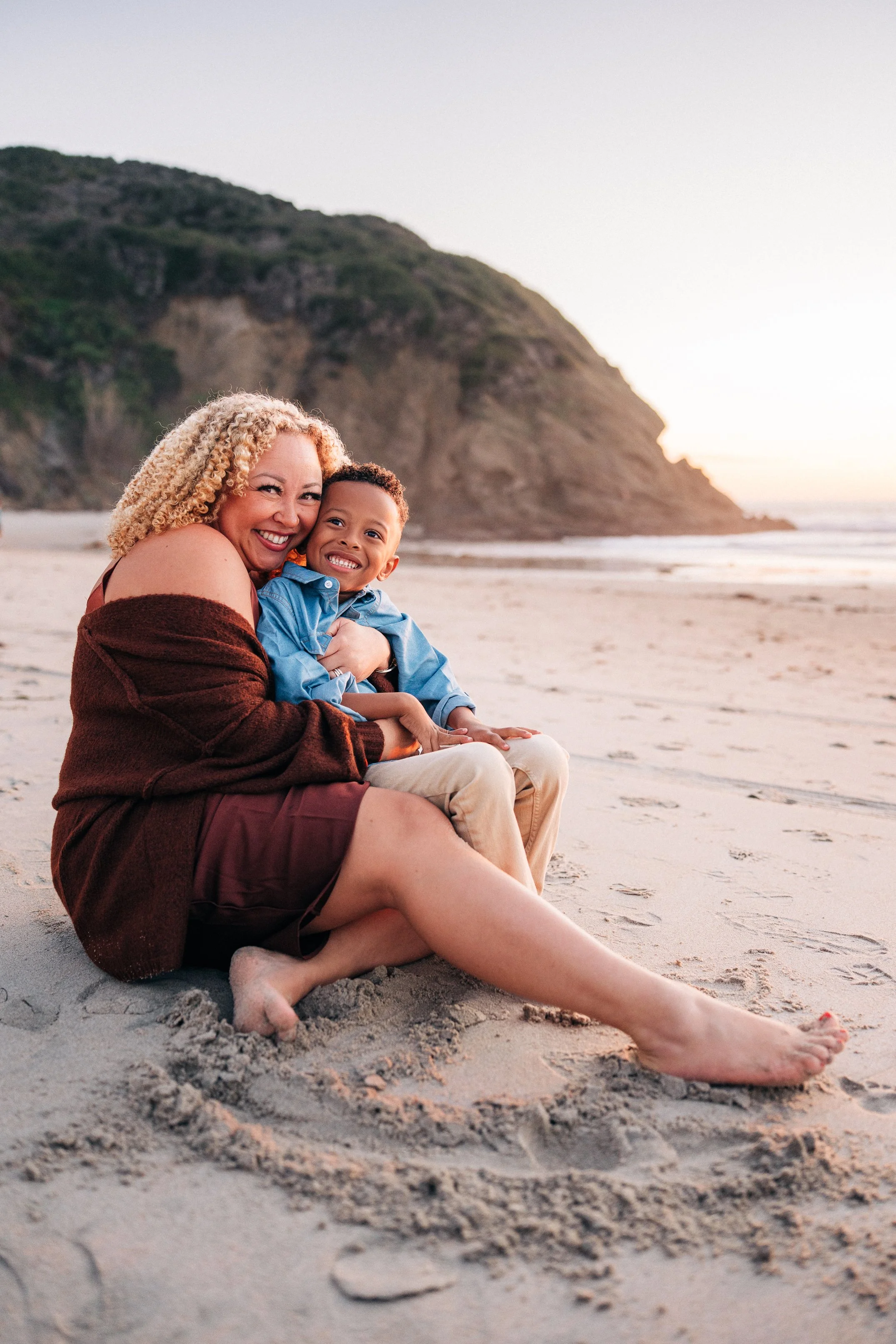 Mom and son sitting together on the sand, smiling at the camera
