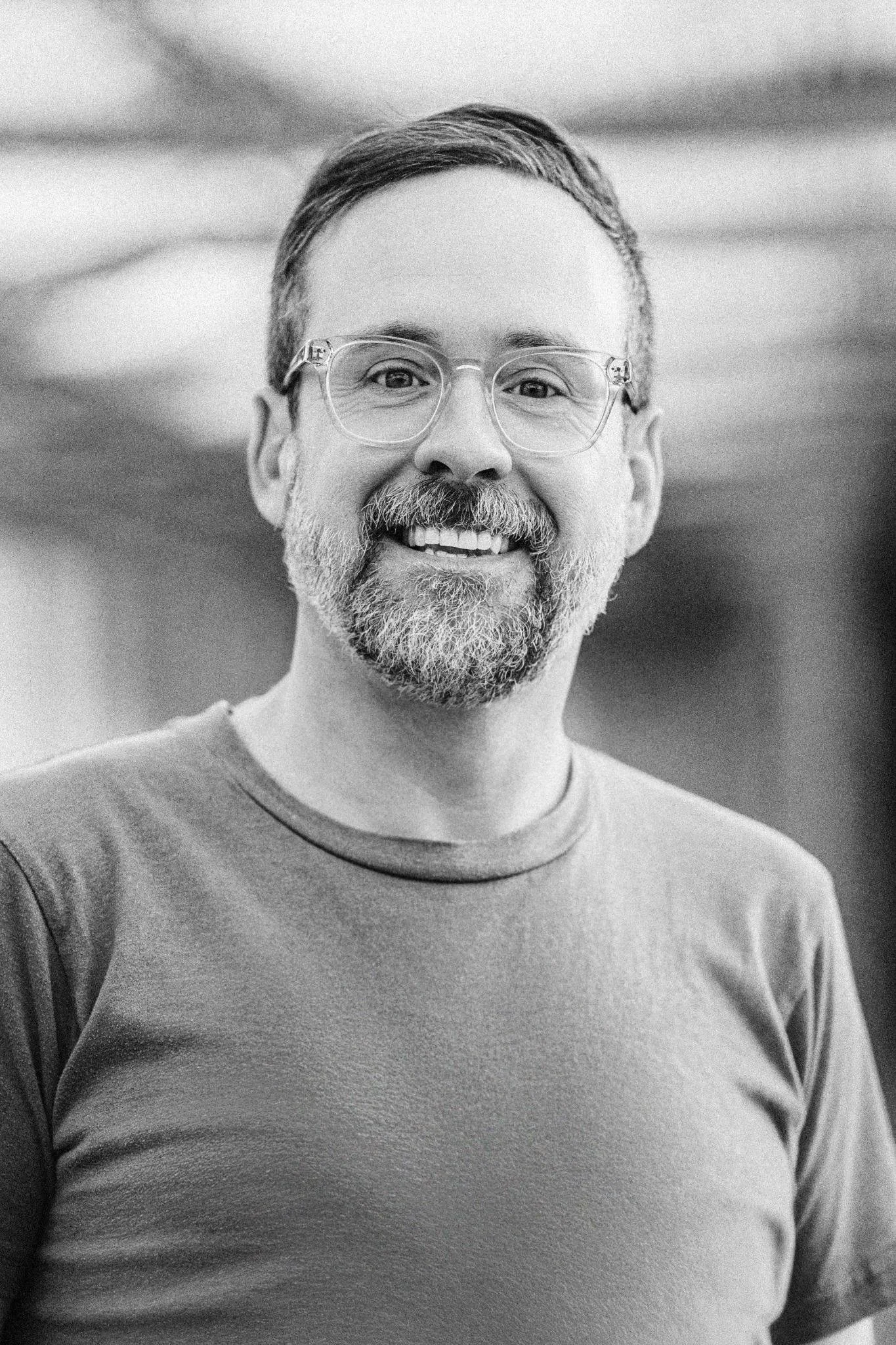 A middle-aged man with glasses, a beard, and short hair, smiling in a close-up black and white portrait.