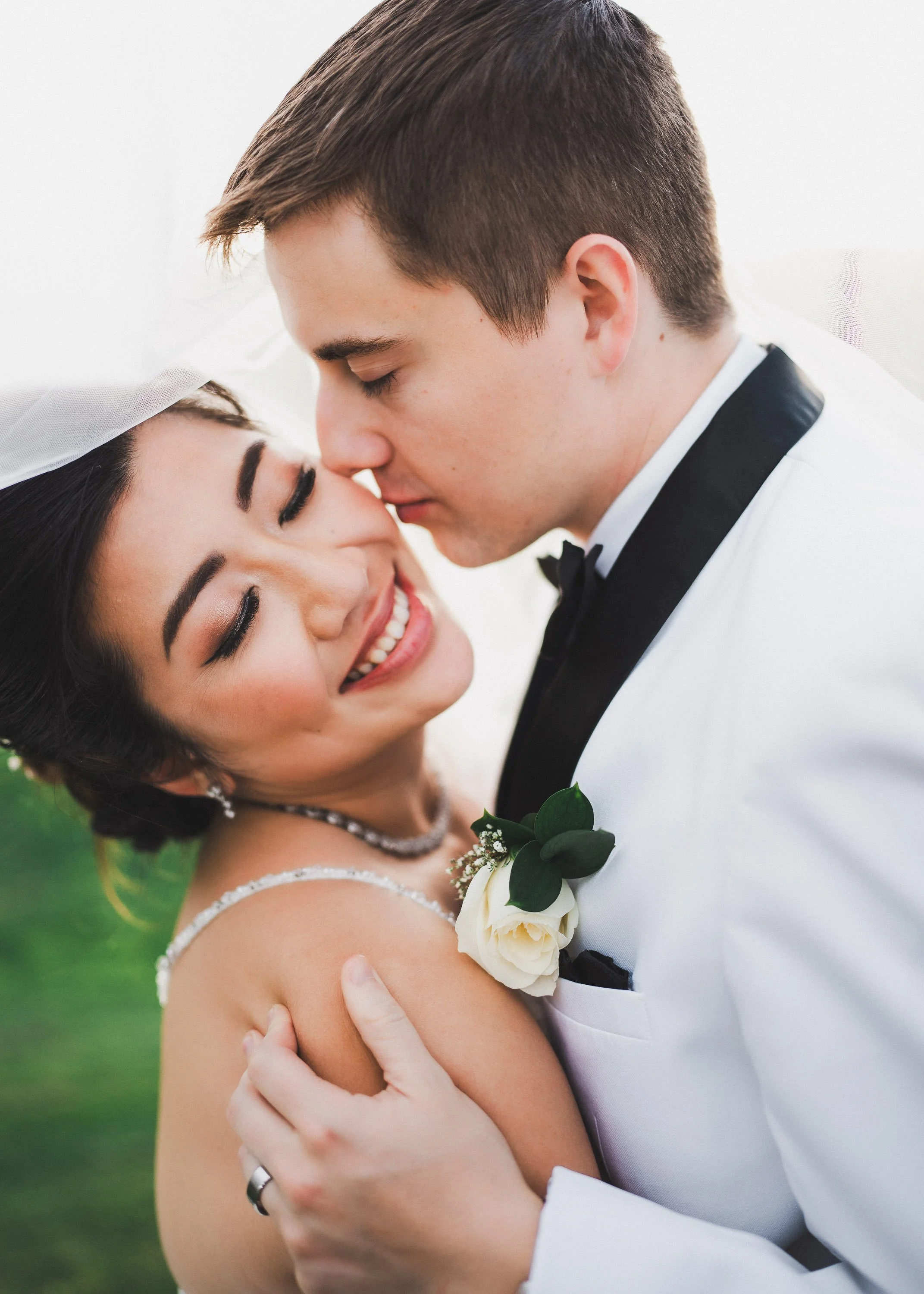 A newly married couple embraces outdoors, with the groom gently holding the bride as they touch foreheads, smiling with eyes closed.