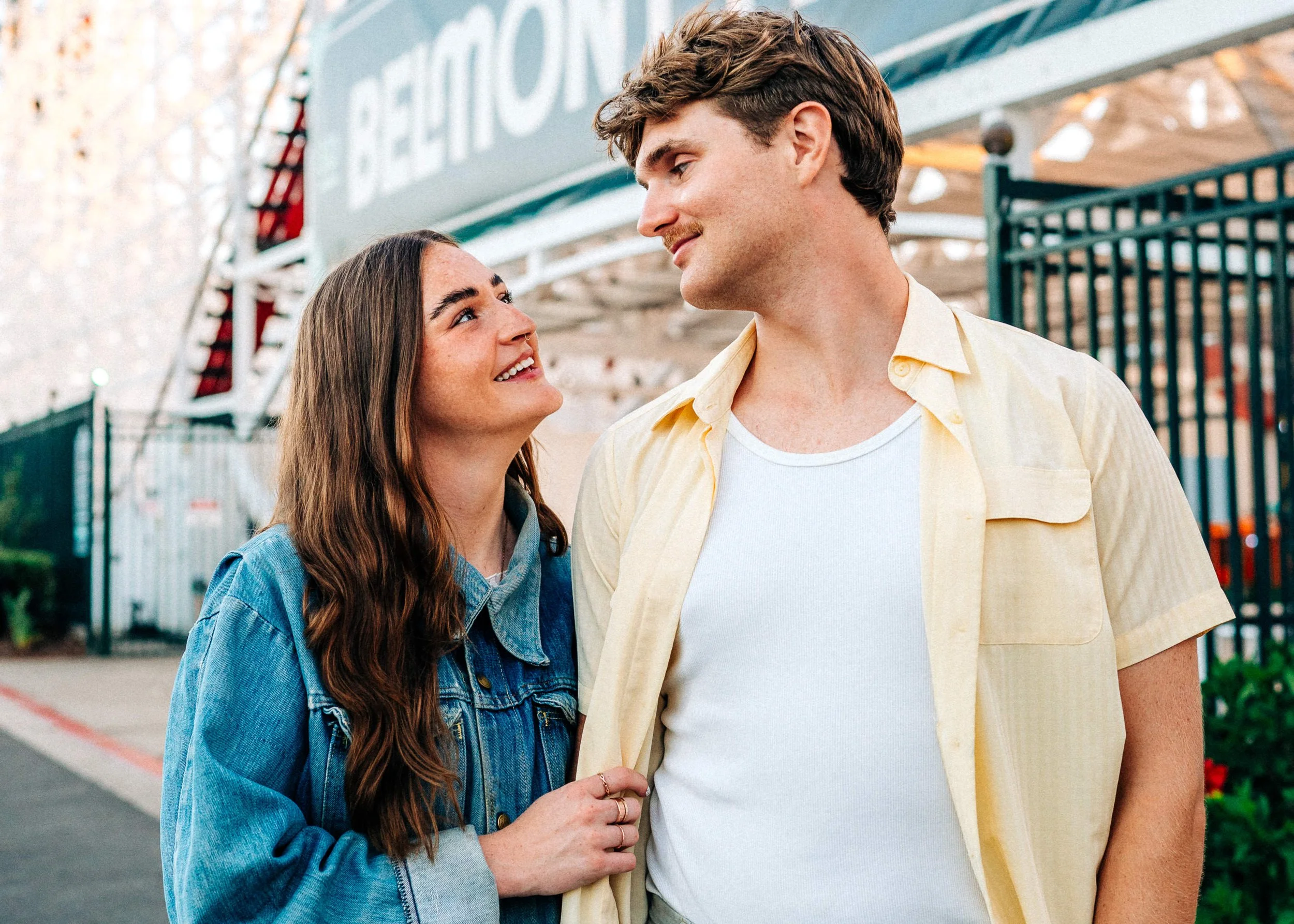 A young woman and man standing close, smiling and looking into each other's eyes outside near a beachside amusement park.