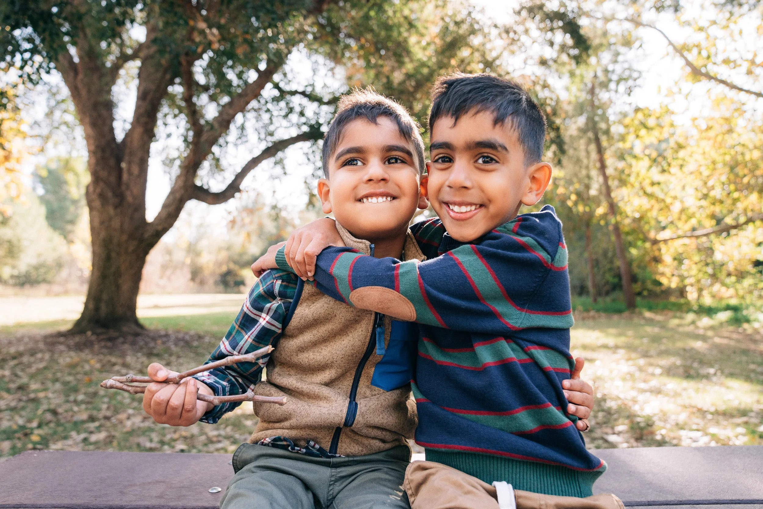 Two young boys happily hugging outdoors in a park with trees and fall foliage in the background.