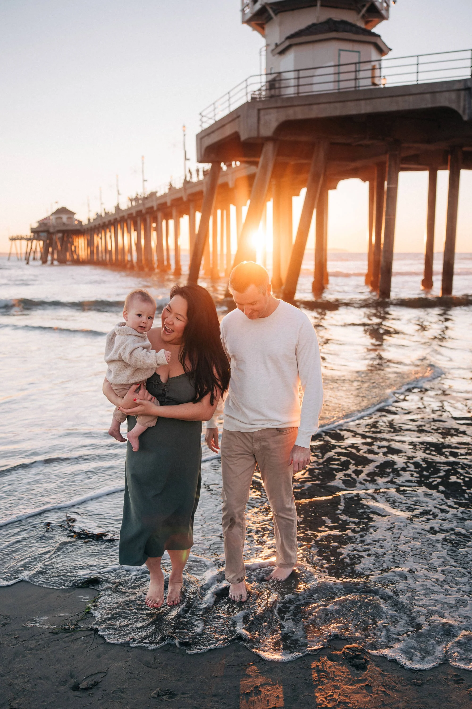 A family of three walking barefoot on the beach at sunset near a pier. The woman is holding a baby and both are smiling, with the man walking beside them.