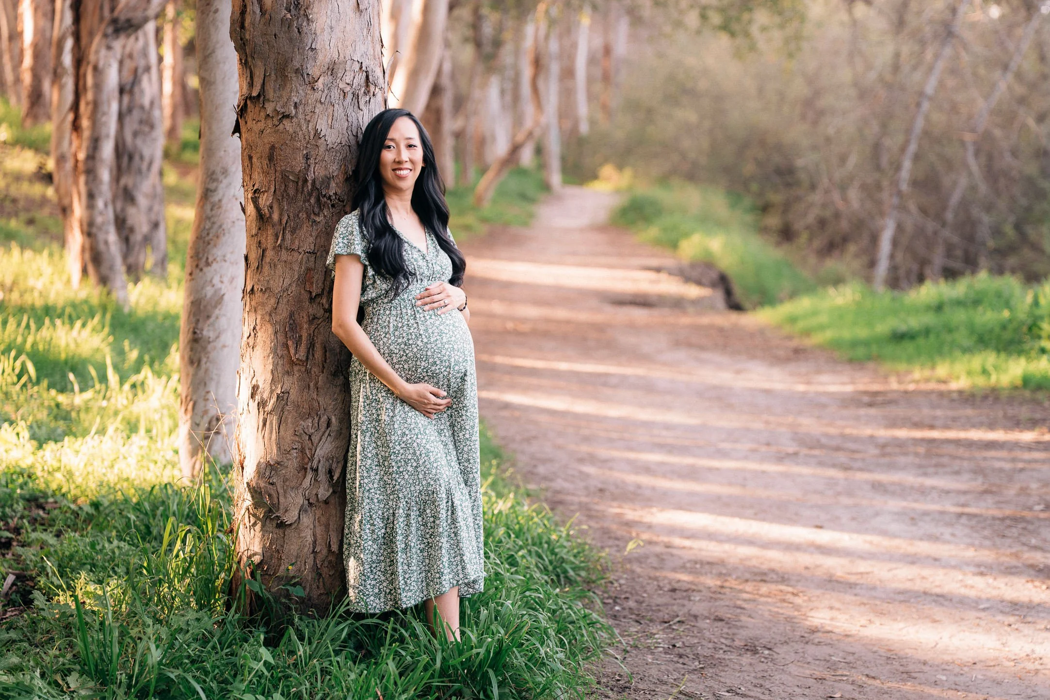 Pregnant woman in floral dress standing next to a tree, smiling, on a dirt path in a forested area during daylight.