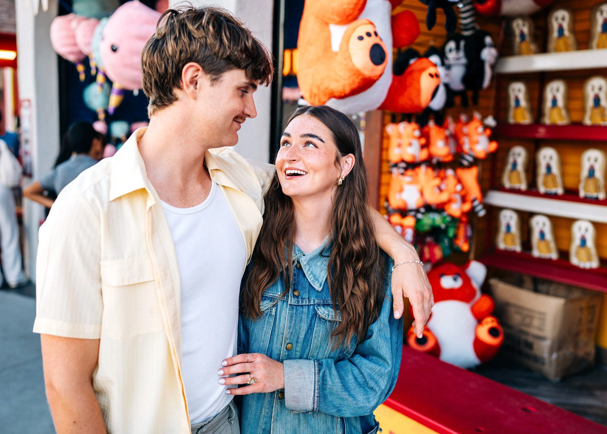 A young couple smiling and looking at each other at a carnival or fair, with plush toys hanging on a wall behind them.