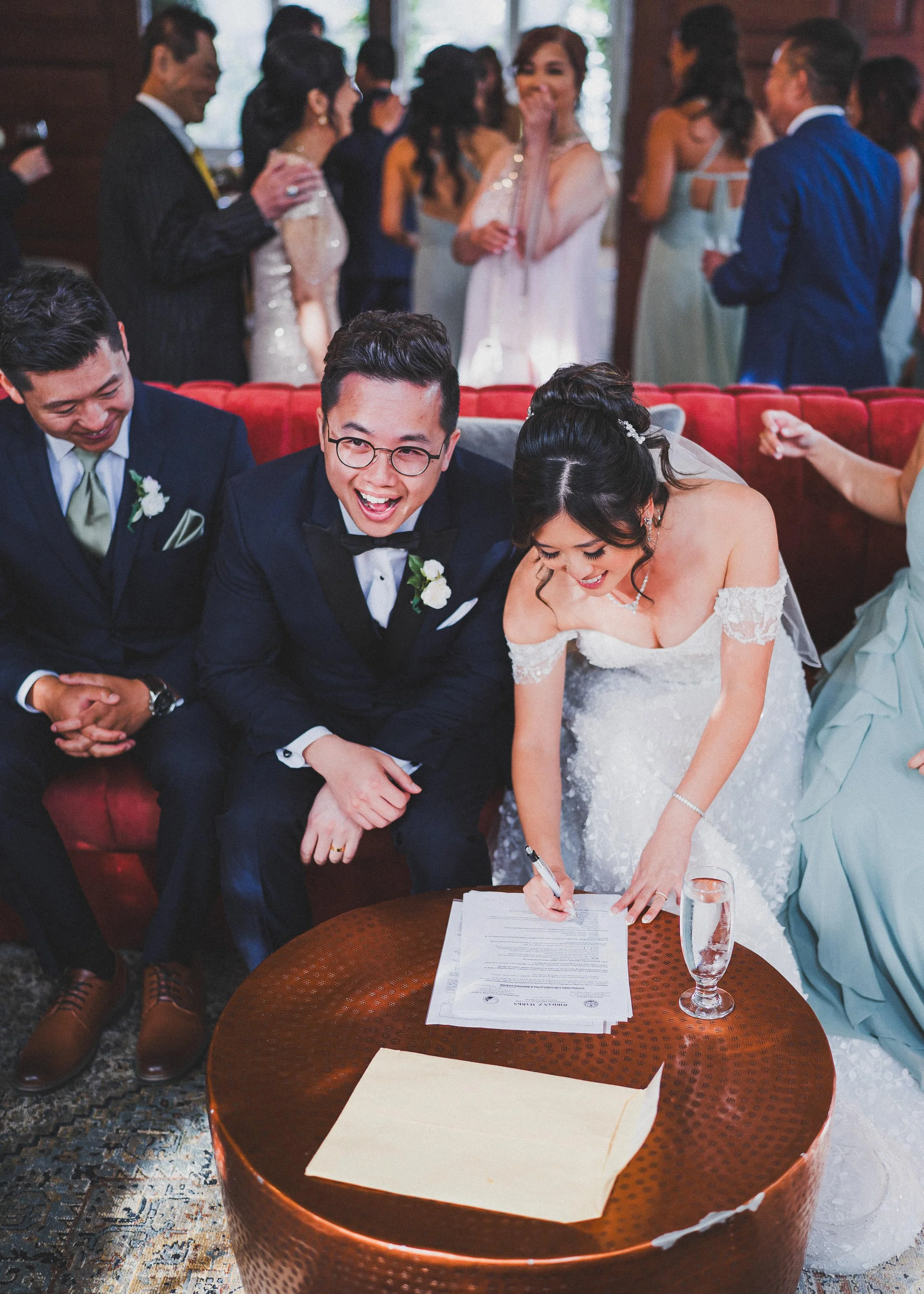 Bride signing marriage documents at her wedding reception, surrounded by groomsmen and wedding guests.