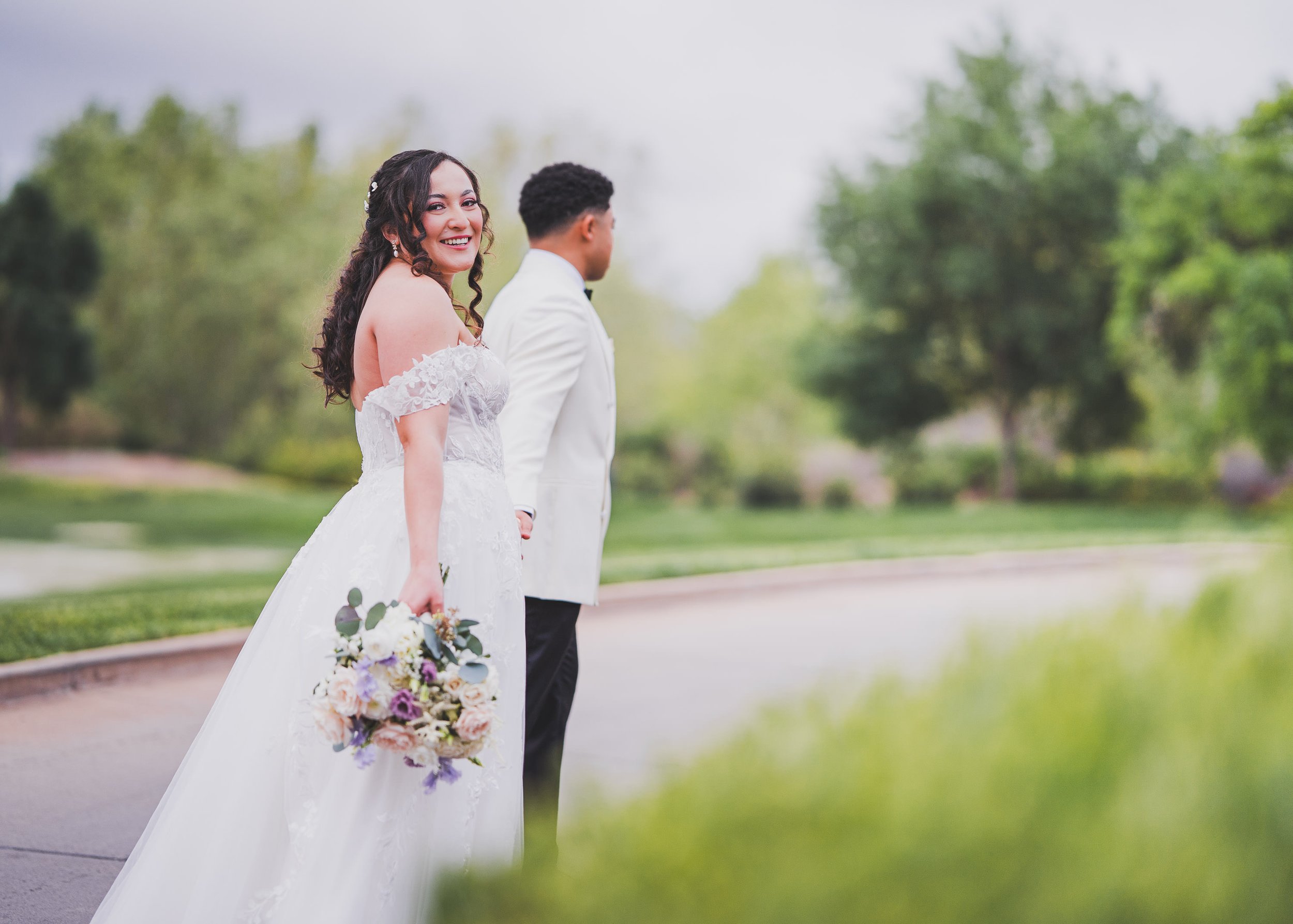 A smiling bride in a white wedding dress holding a bouquet of flowers, standing outdoors with a groom in a white suit, blurred green trees in the background.