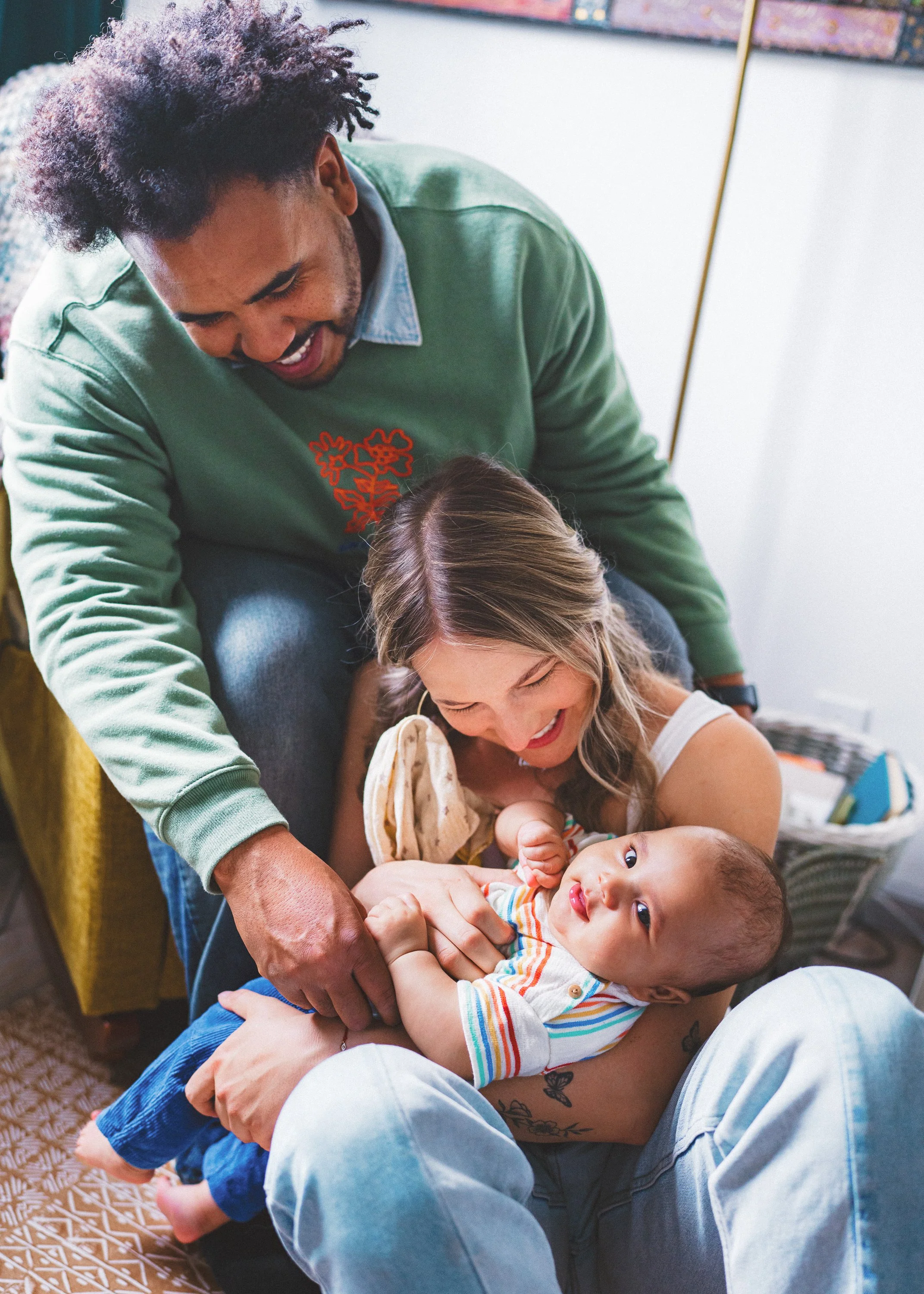 A smiling woman holding a baby with a striped shirt on her lap, while a man in a green sweatshirt interacts with them, all in a cozy indoor setting.