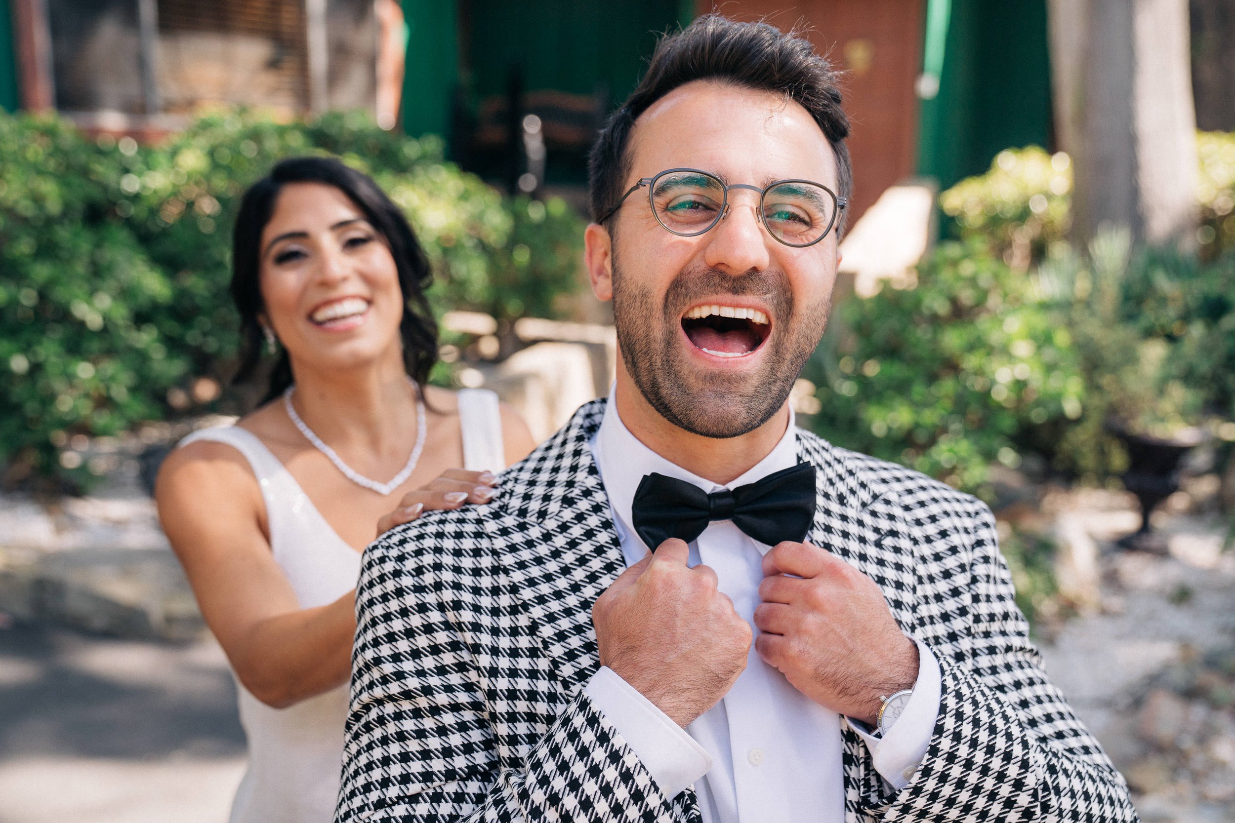 A man wearing glasses and a checkered jacket with a bow tie enjoying a moment with a woman in a white dress behind him, outdoors surrounded by greenery.