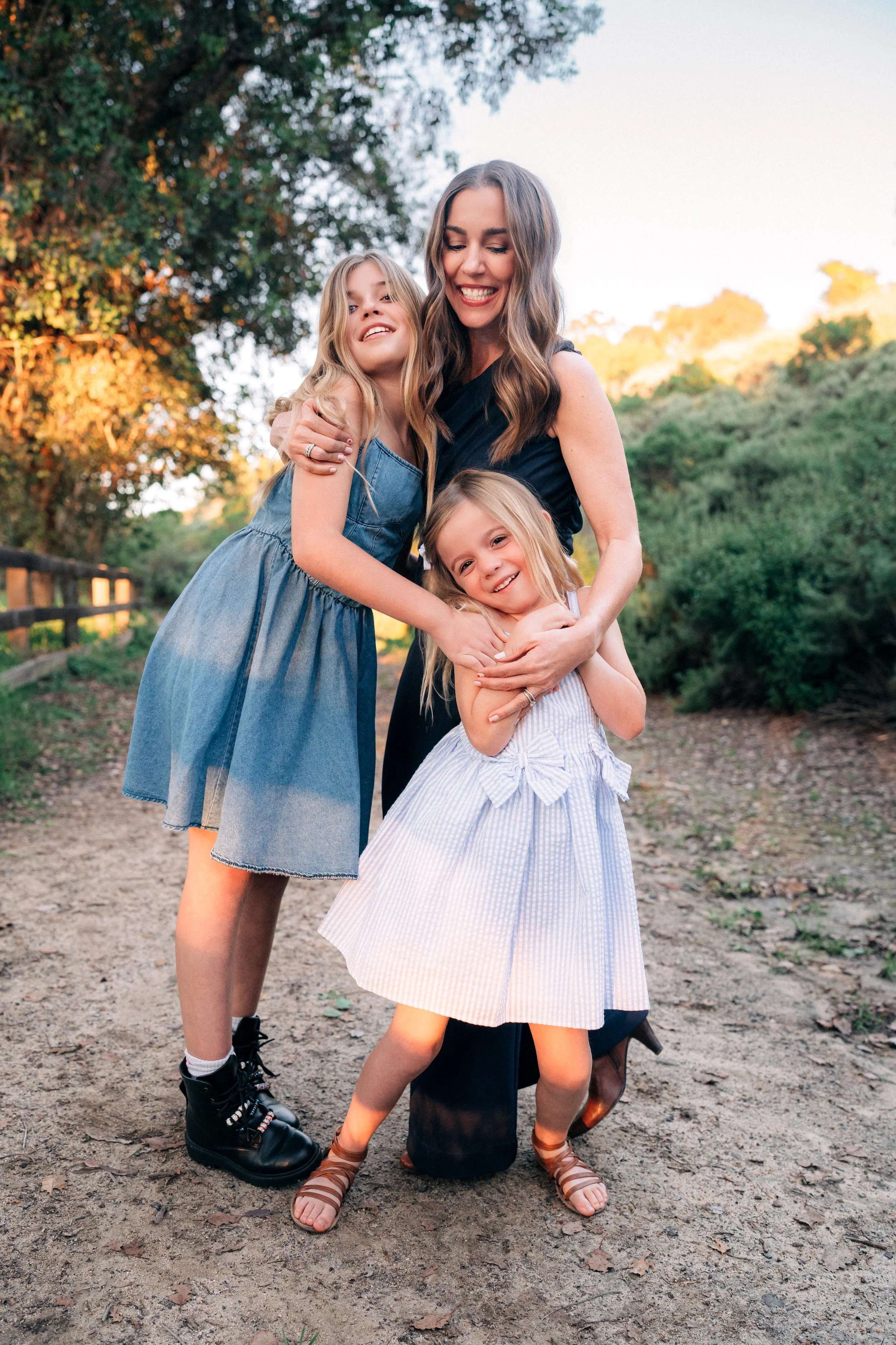 A woman with two young girls hugging outdoors during sunset, surrounded by trees and greenery.