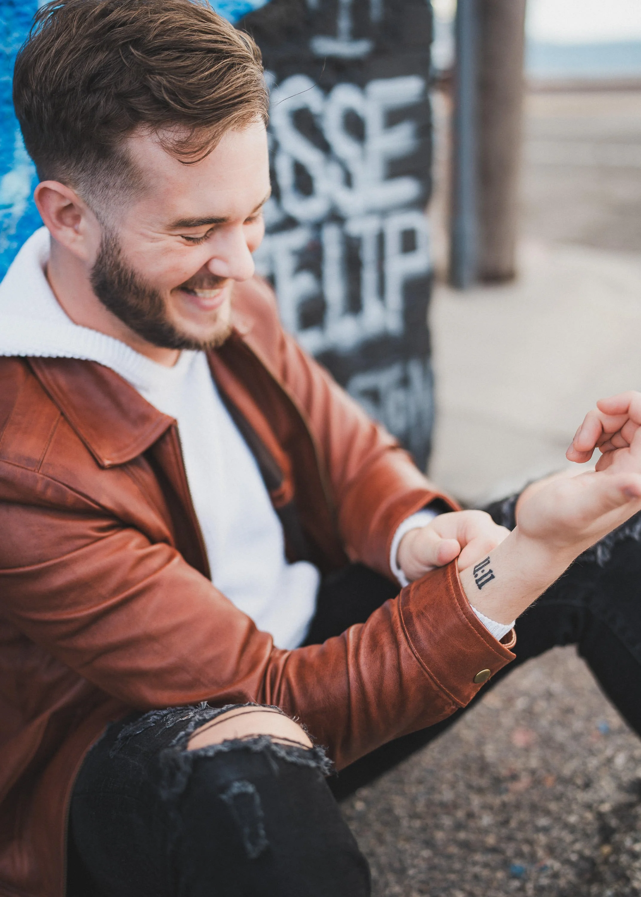 A young man with a beard, wearing a white shirt and brown leather jacket, sitting on the ground and smiling while looking at his phone. He has a tattooed wrist that says 'ill' and is wearing ripped black jeans. In the background, there is a chalkboar