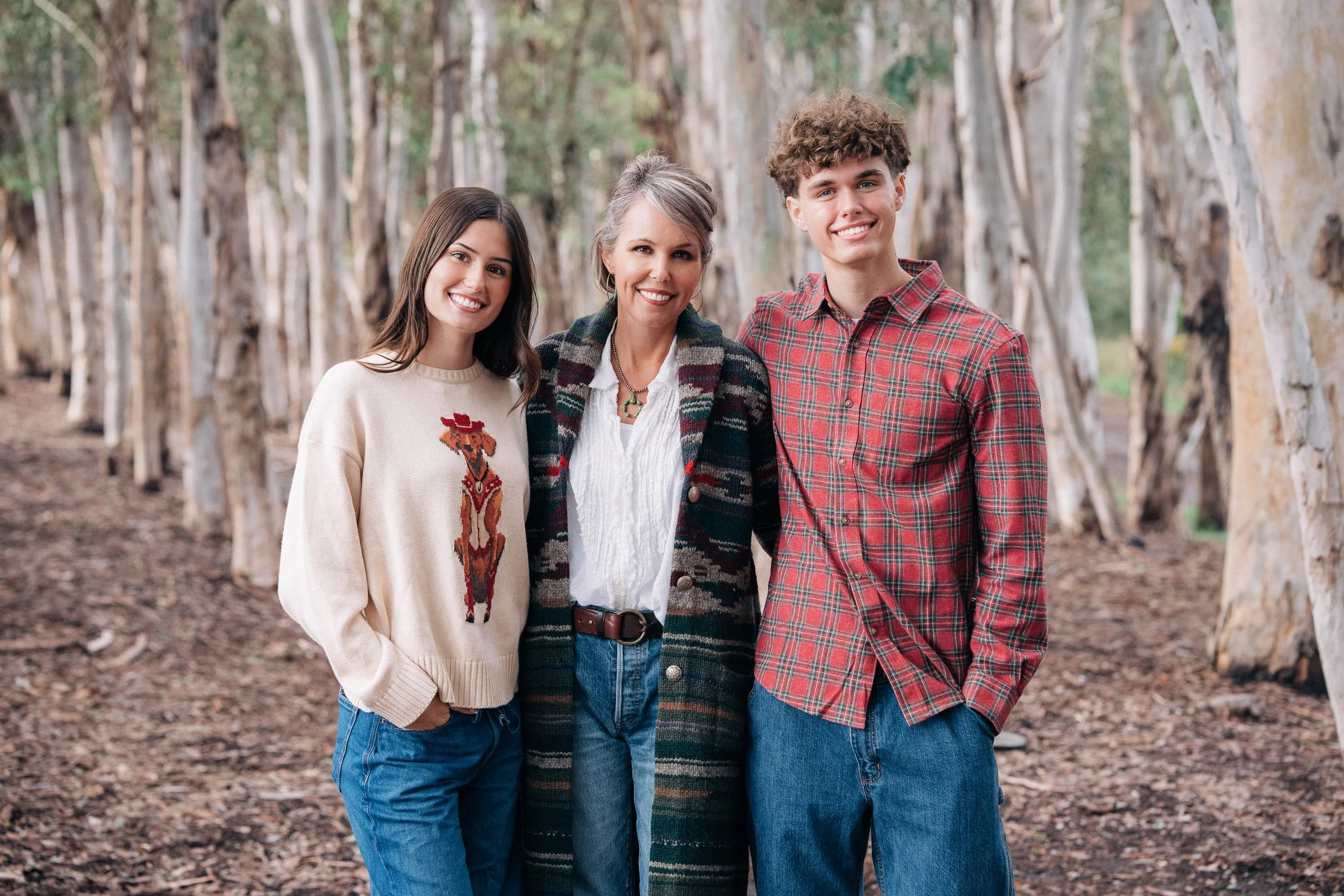 A woman and two teenagers standing outdoors on a dirt path surrounded by tall trees with peeling bark, smiling at the camera.