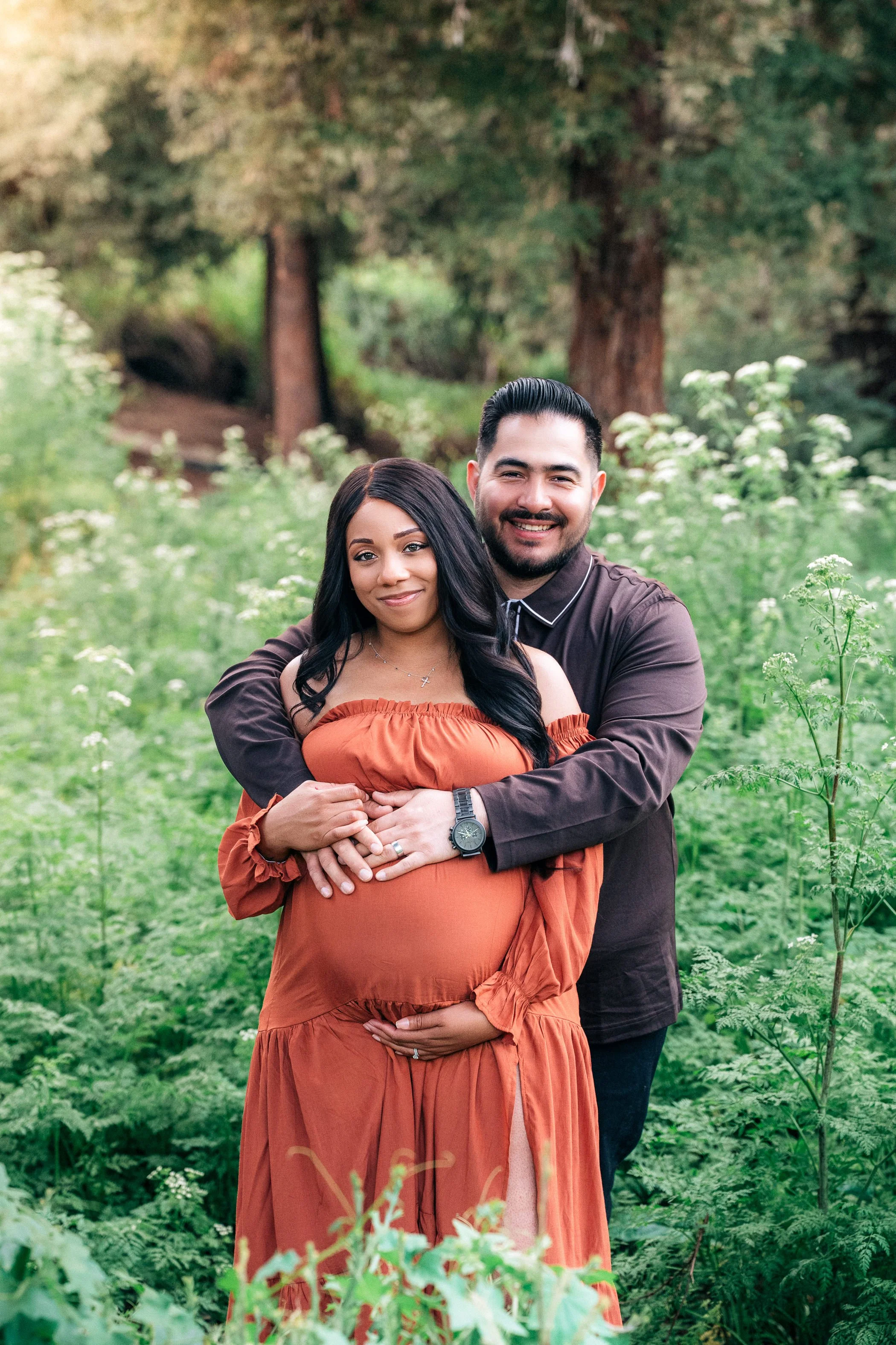 A pregnant woman and her partner embracing outdoors in a green, wooded area.