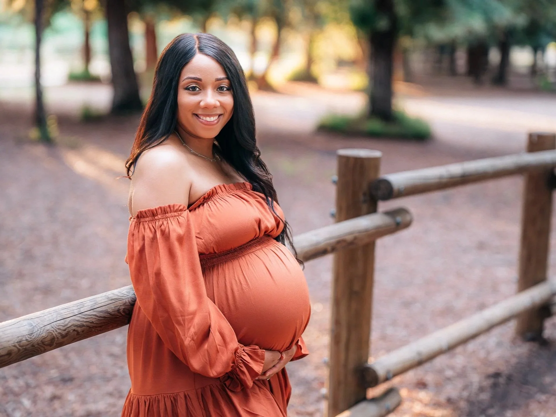 A pregnant woman with long dark hair smiling outdoors in a park with trees, standing by a wooden fence, wearing an off-the-shoulder burnt orange dress.