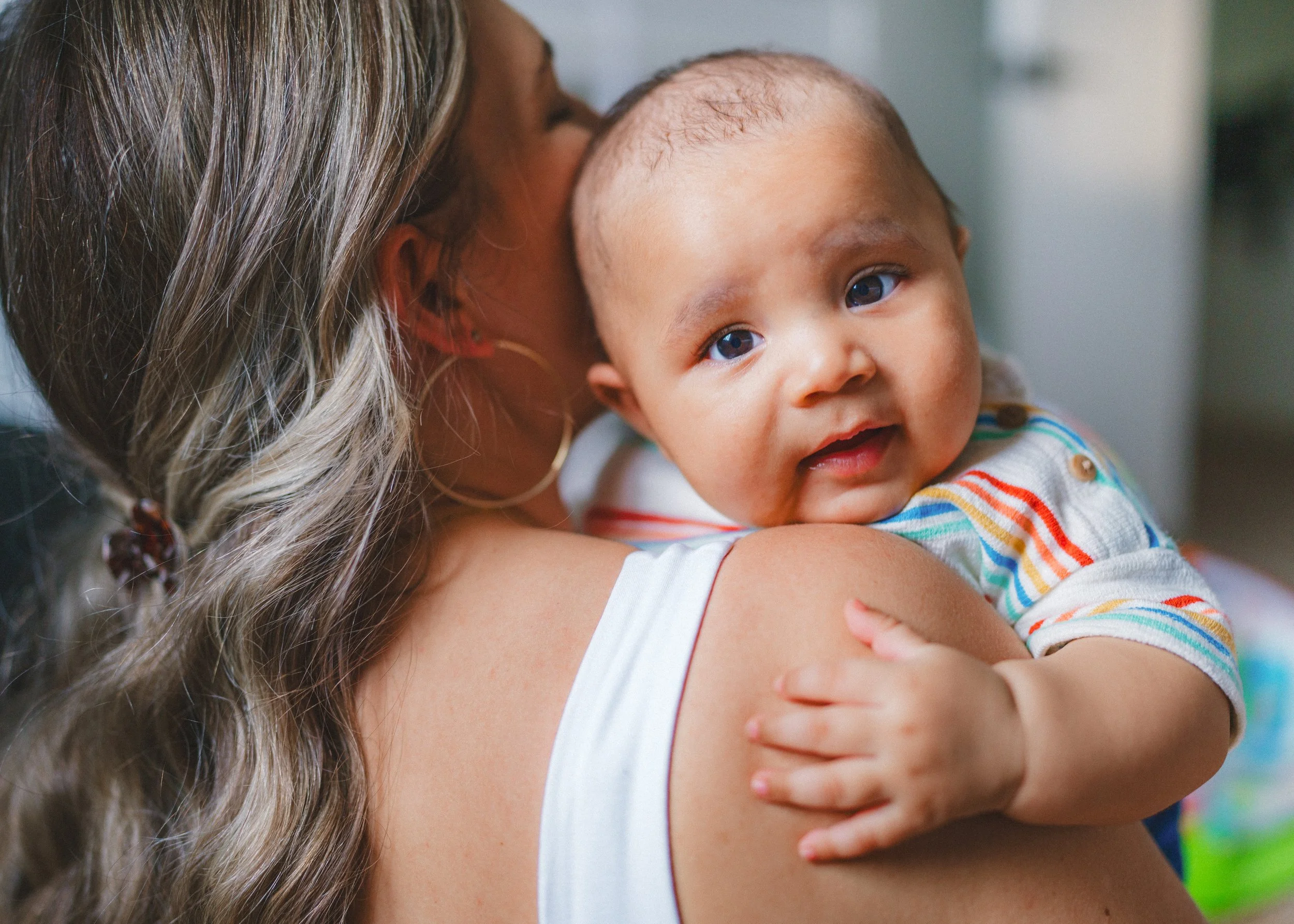 A woman holding a baby with light skin, blue eyes, and short hair. The woman’s face is partially visible, and she has long, wavy hair with highlights and a large hoop earring. The baby is resting on the woman’s shoulder, looking at the camera, wearin