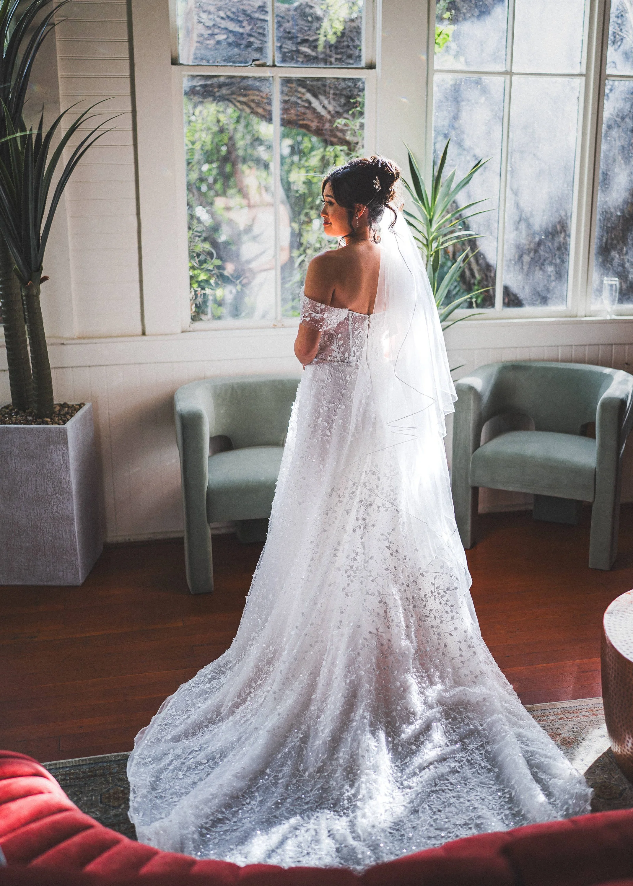 A bride in a white off-shoulder wedding dress and veil stands in a sunlit room with large windows, green chairs, potted plants, and wooden floors.
