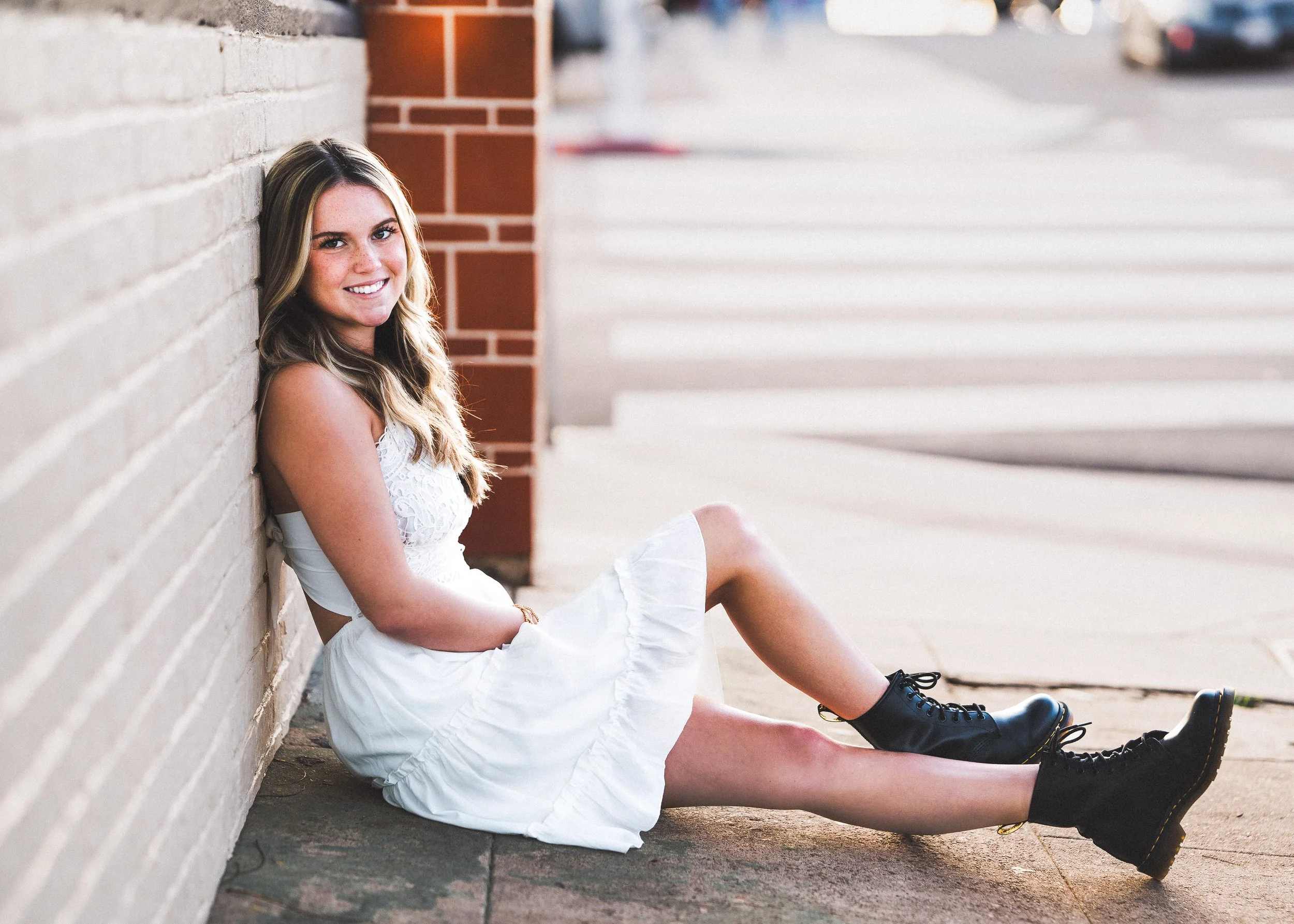A young woman with long, wavy hair sitting on the sidewalk, leaning against a brick wall, smiling at the camera, wearing a white dress and black combat boots.