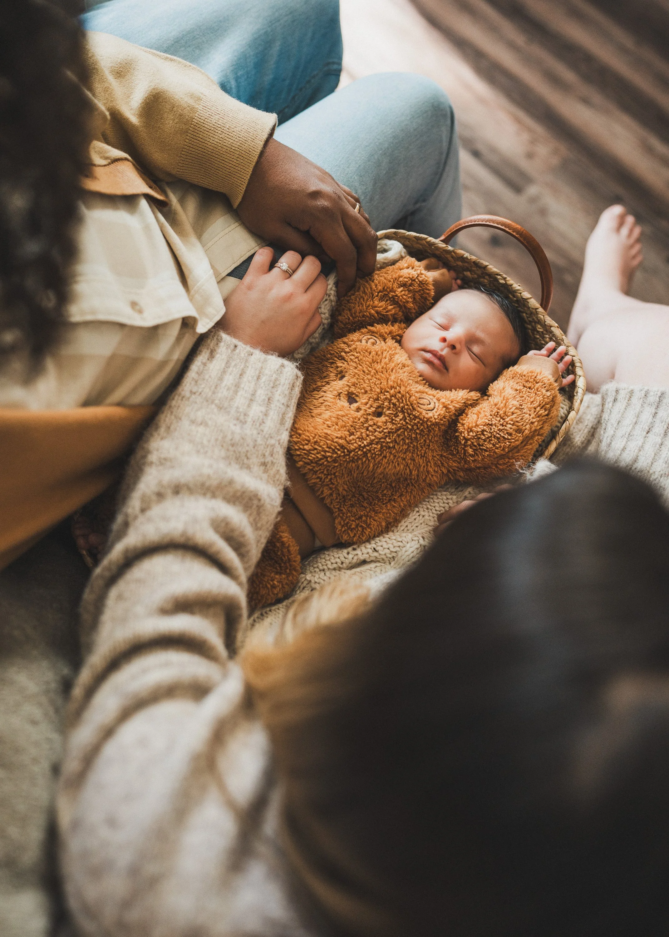 A newborn baby sleeping in a brown fuzzy sweater in a woven basket, surrounded by adults with layered clothing, on a wooden floor.