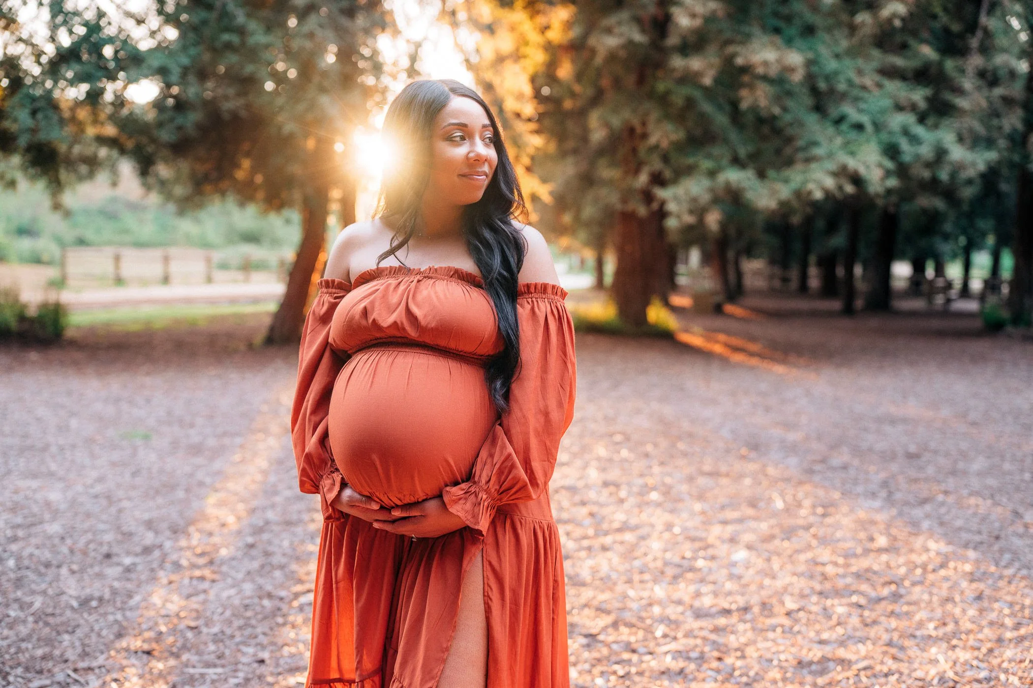 Pregnant woman in an orange dress holding her belly outdoors during sunset, with trees and a path in the background.
