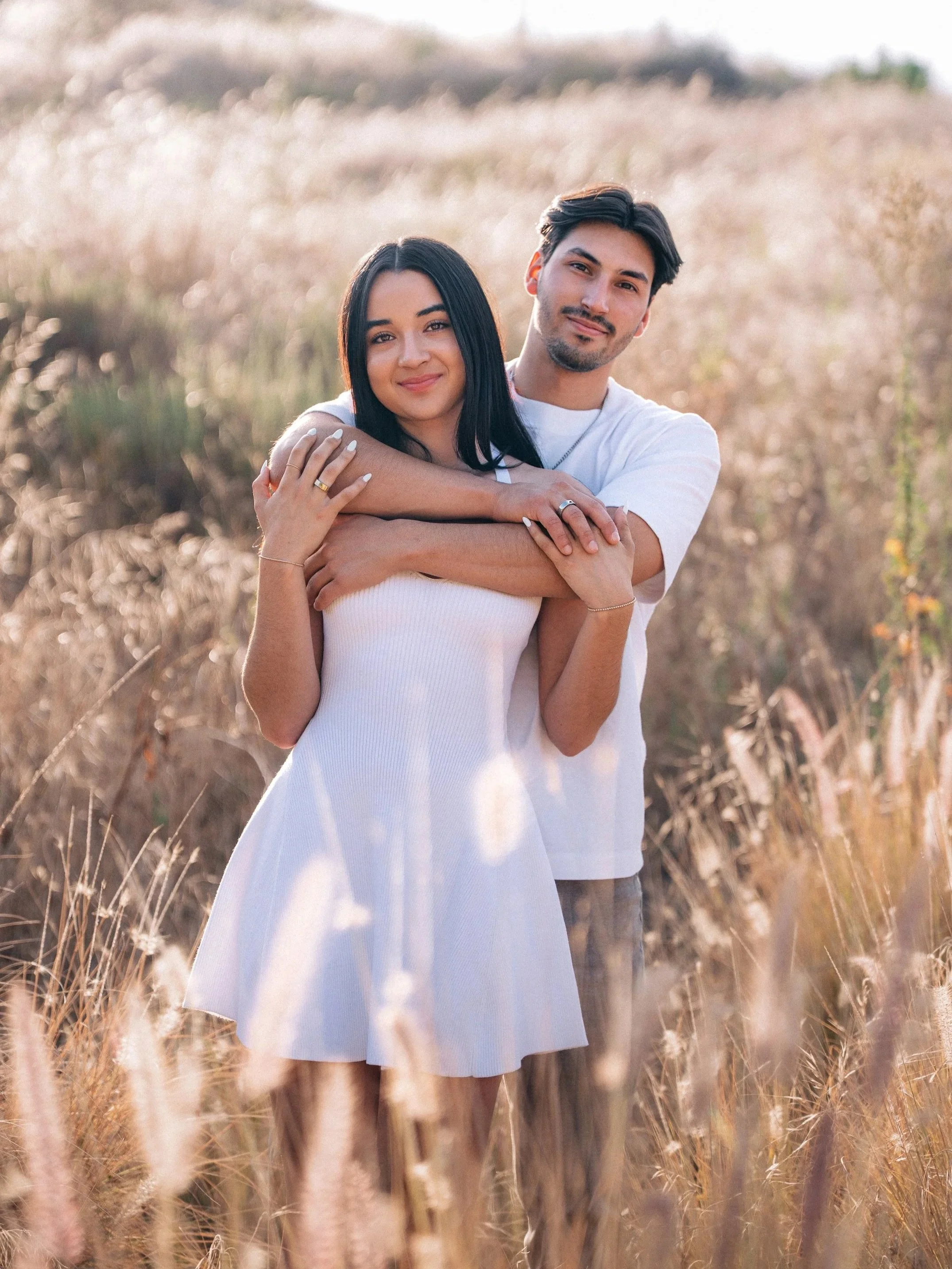 A young couple standing in a field of tall, dry grass during sunset, with the woman leaning into the man's embrace, both smiling softly at the camera.