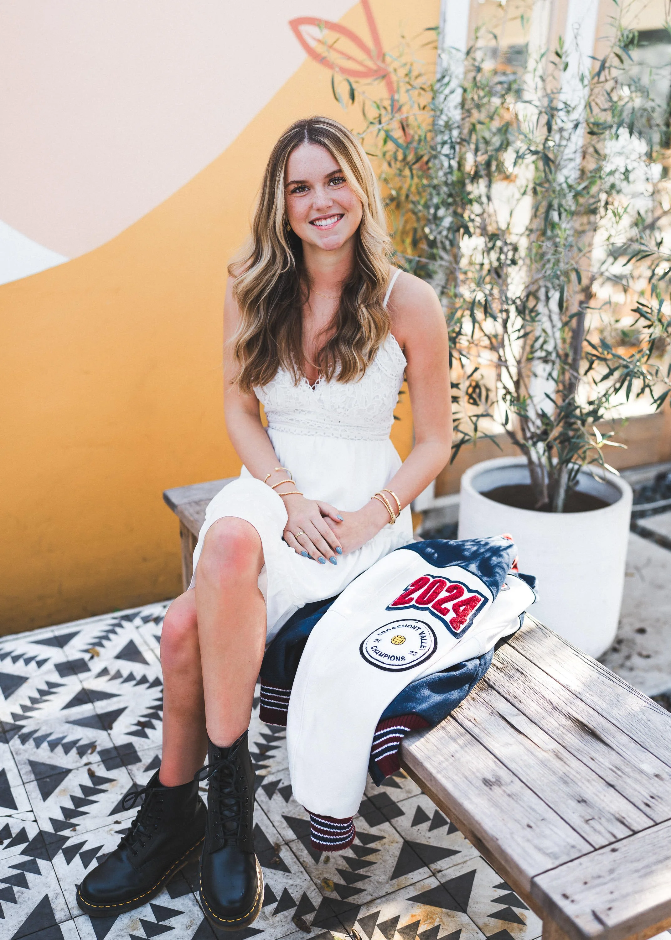 Young woman with long wavy hair smiling, sitting on a wooden bench outdoors, wearing a white dress and black combat boots, with a jacket that has a 2024 patch and a logo, next to a large potted plant and colorful wall art.