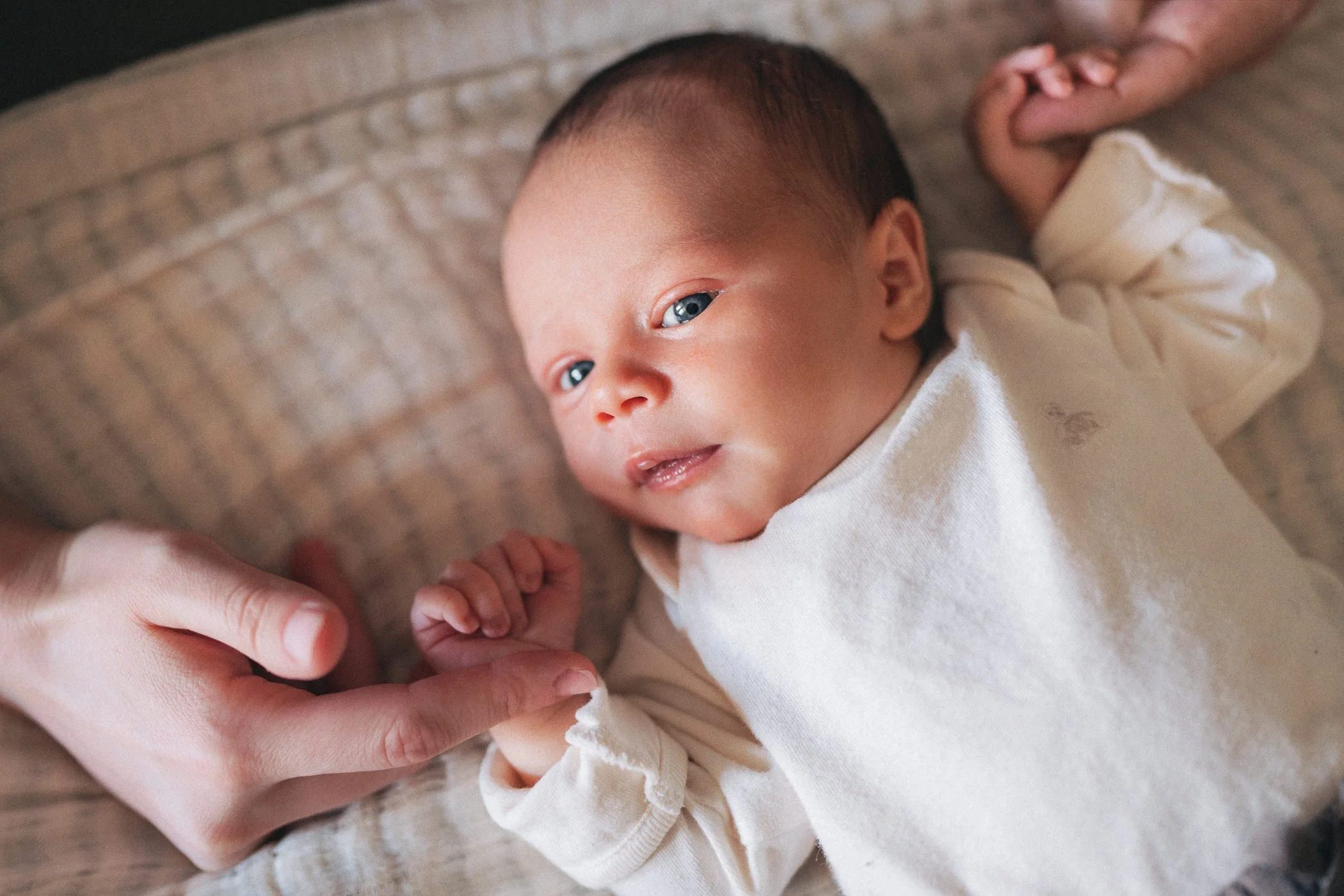 A newborn baby lying on a textured beige blanket, looking at the camera while holding an adult's finger.