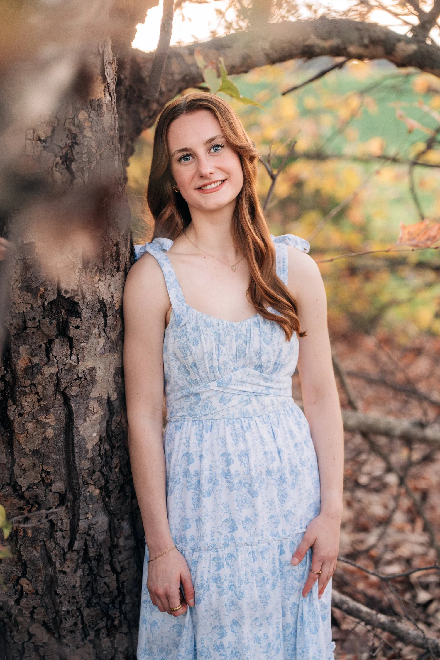 Senior portrait of Emily leaning against a tree at golden hour in a blue floral dress, Coto de Caza