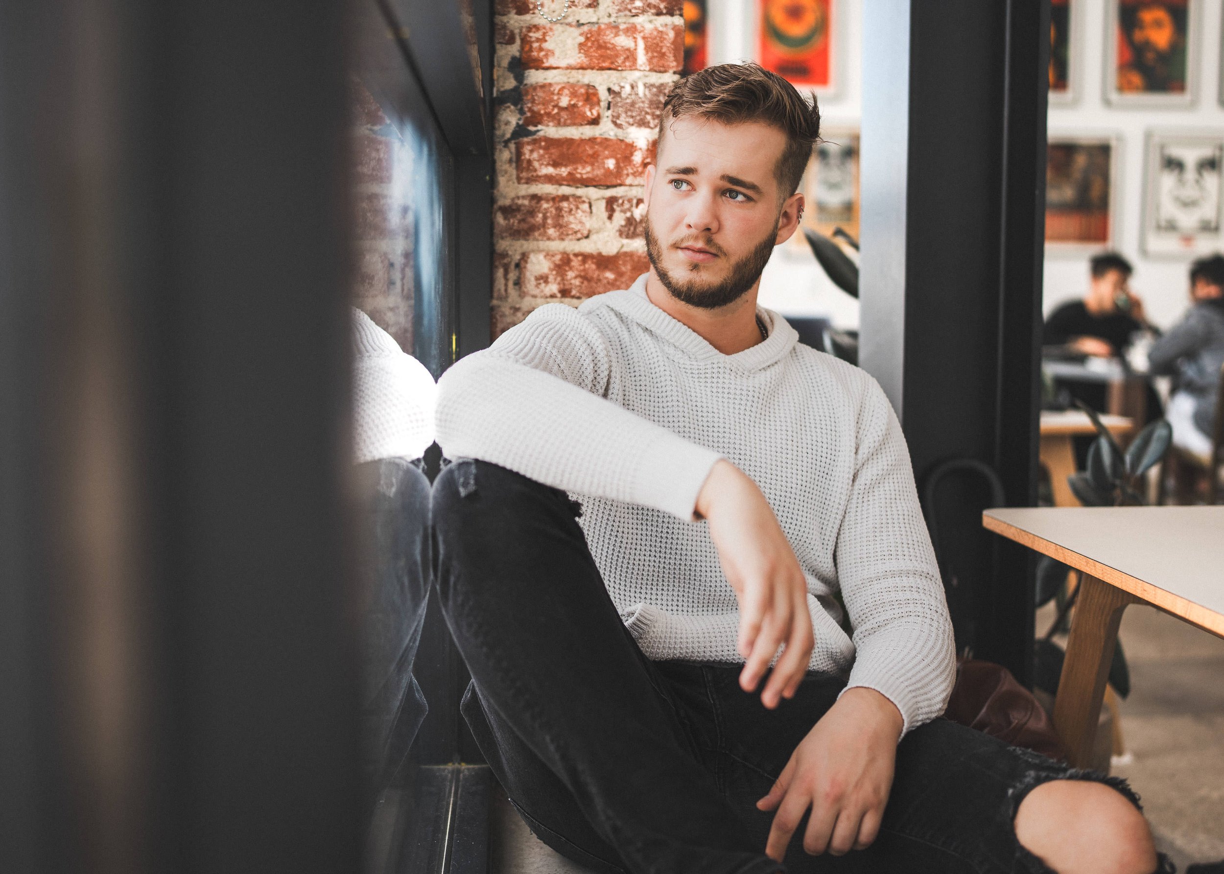 A young man with a beard, wearing a white sweater and black pants, sitting in a cafe near a window, looking thoughtfully to the side.