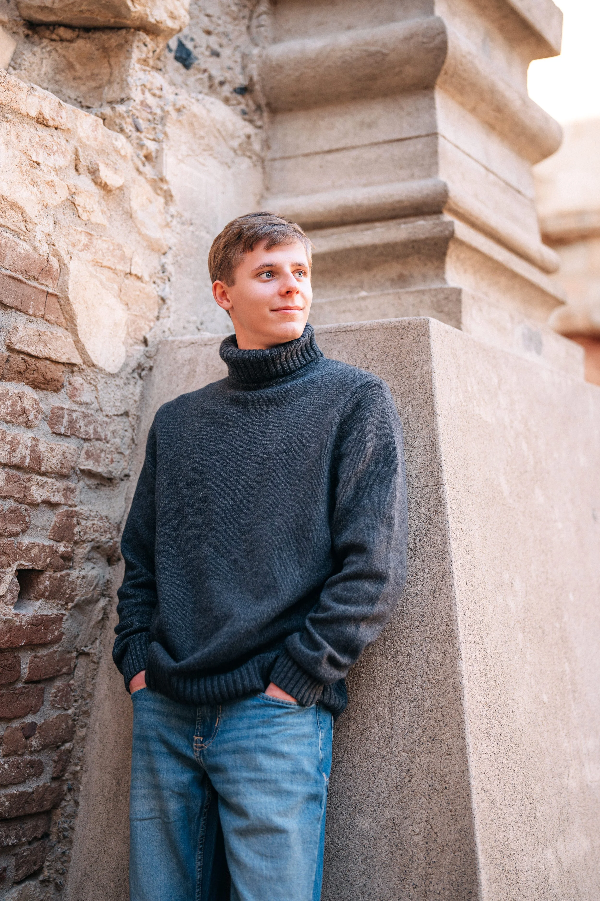 A young man in a dark gray turtleneck sweater and blue jeans leaning against a stone wall and pillar outside during daytime.
