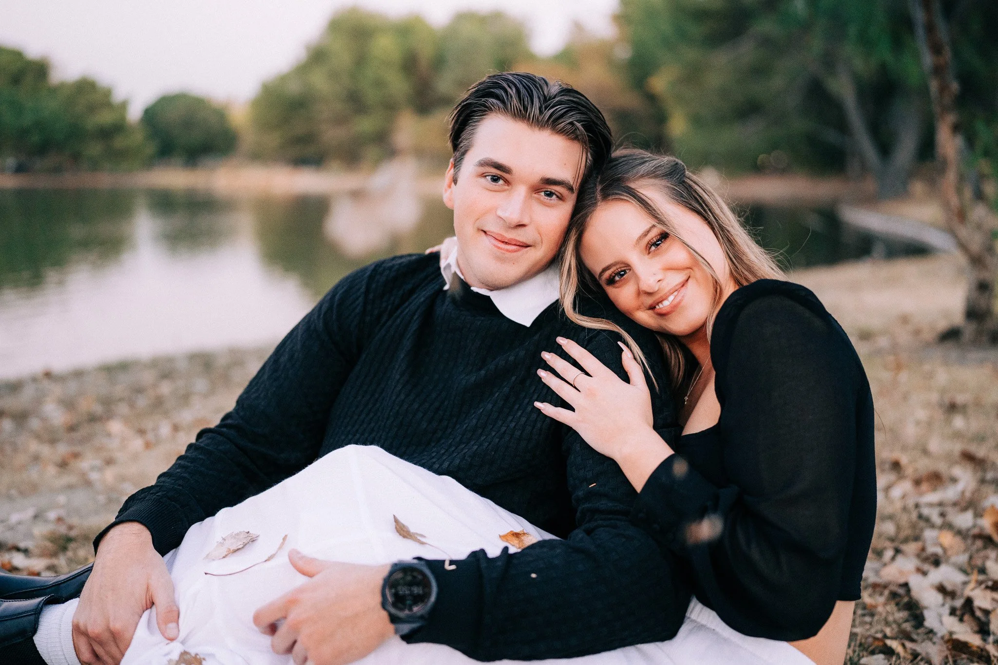 A smiling young couple sitting on the ground near a lake, leaning into each other, with trees in the background during autumn with fallen leaves.