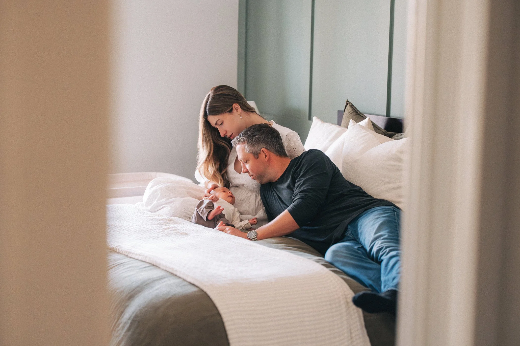 A family of three, a woman, a man, and a baby, sitting on a bed in a bedroom. The woman is holding the baby, and the man is interacting with the baby on the bed.