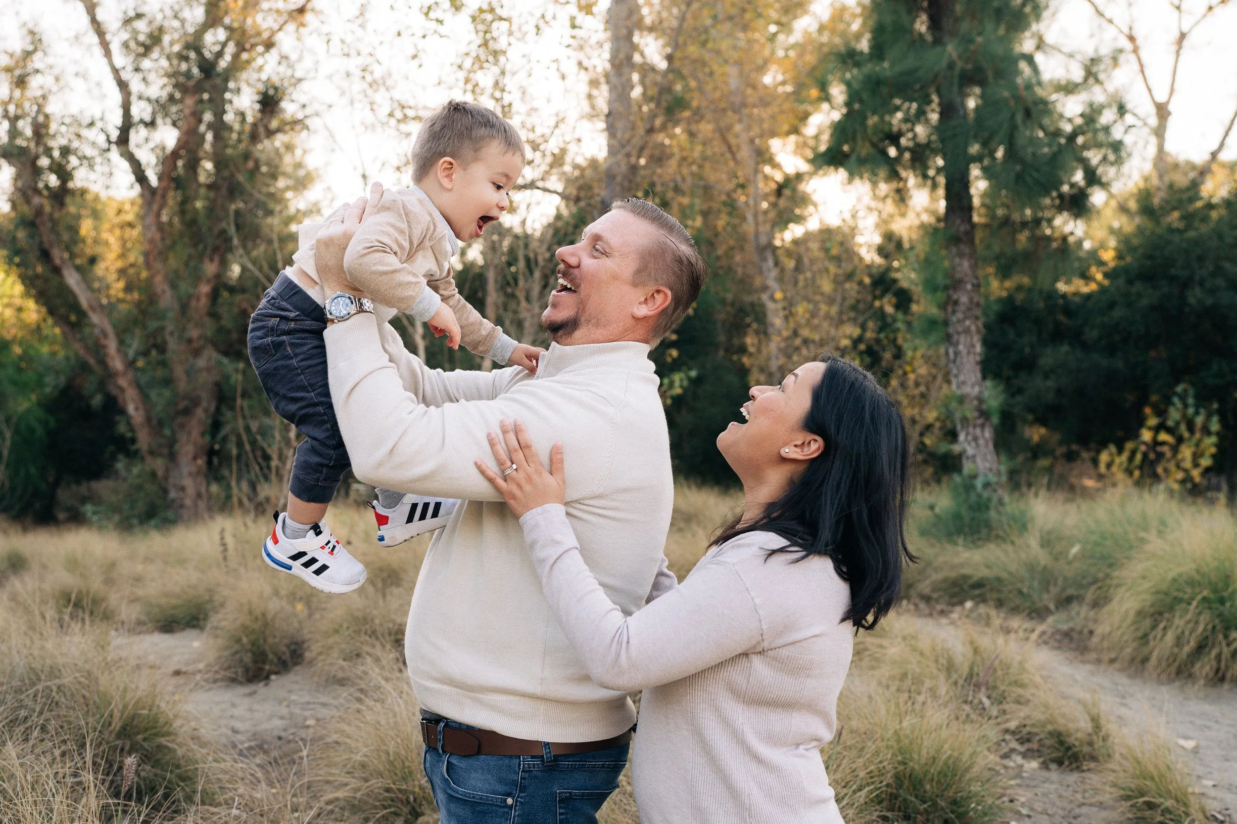 A family of three, a father, mother, and young son, enjoying an outdoor moment in a park during autumn. The father is lifting the boy into the air while the mother looks up smiling.