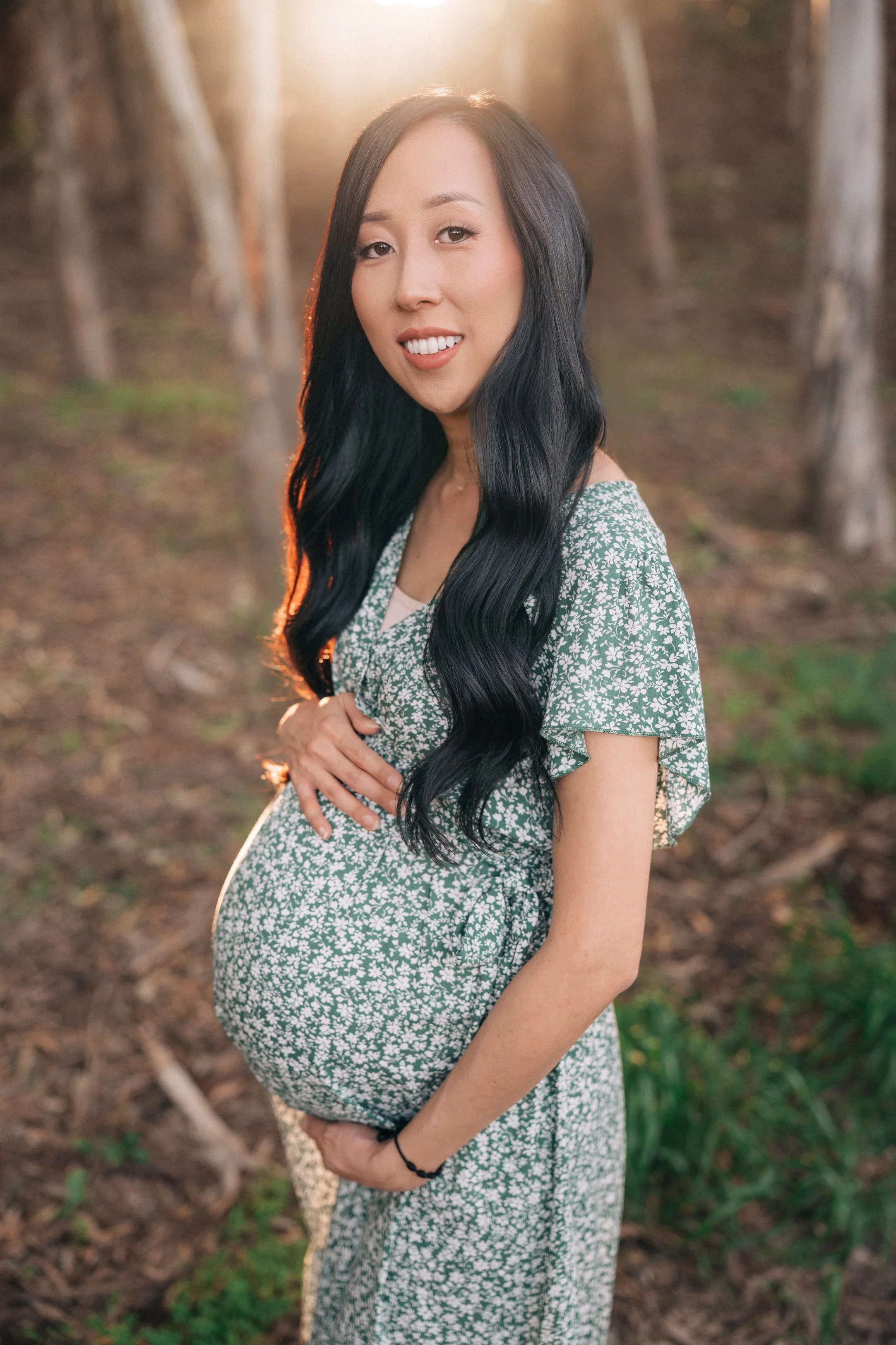 A pregnant woman with long black hair, smiling, standing in a forested area during sunset, wearing a green dress with white floral pattern.