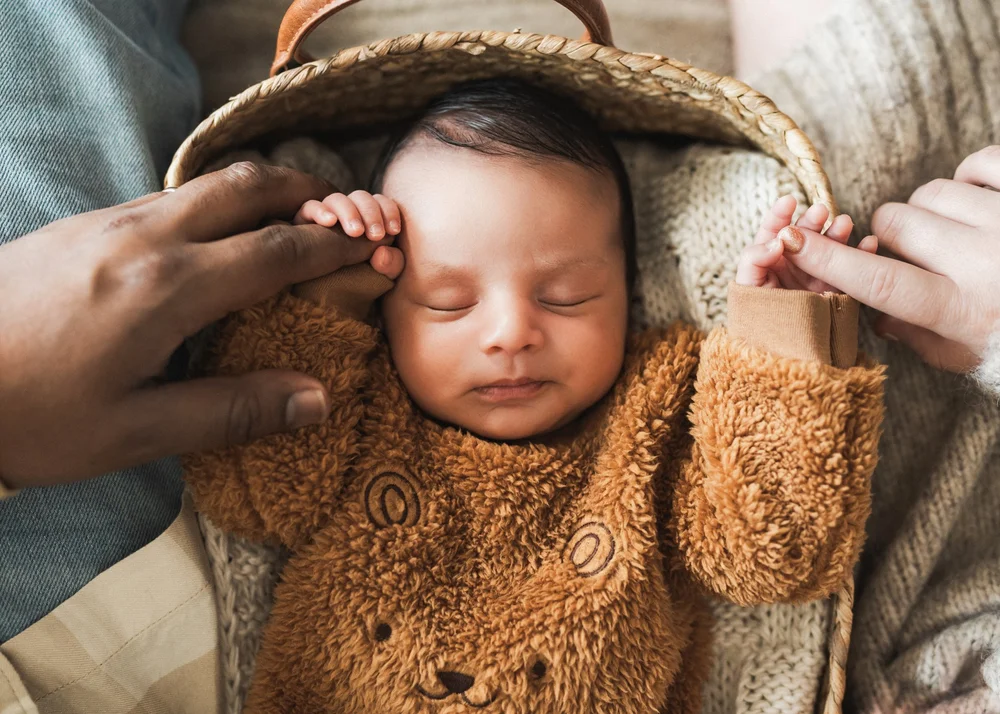 Close-up of newborn sleeping in teddy bear outfit with both parents' hands gently framing the basket, Nathan and Ana in-home session San Diego