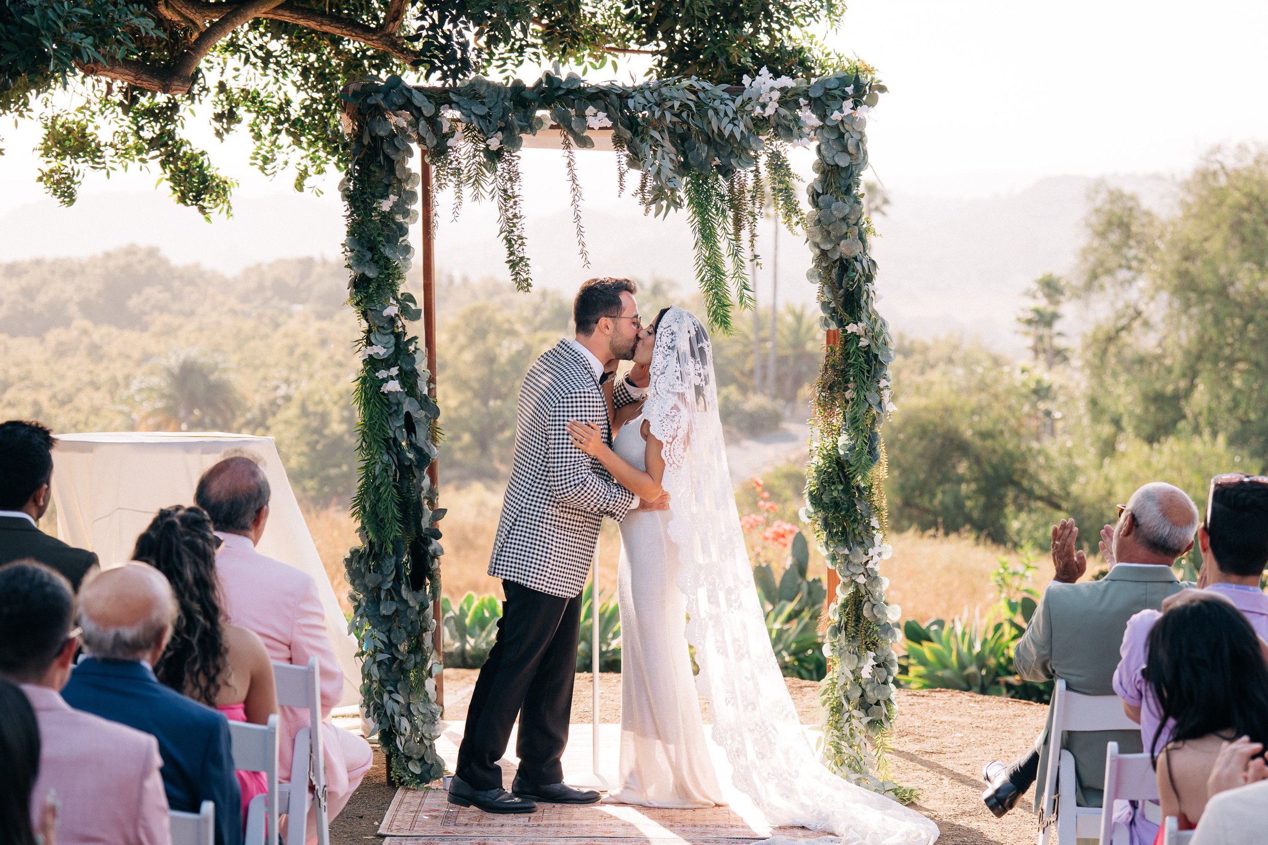 A wedding ceremony outdoors with a bride and groom kissing under a floral arch, surrounded by seated guests, in a natural scenic setting.