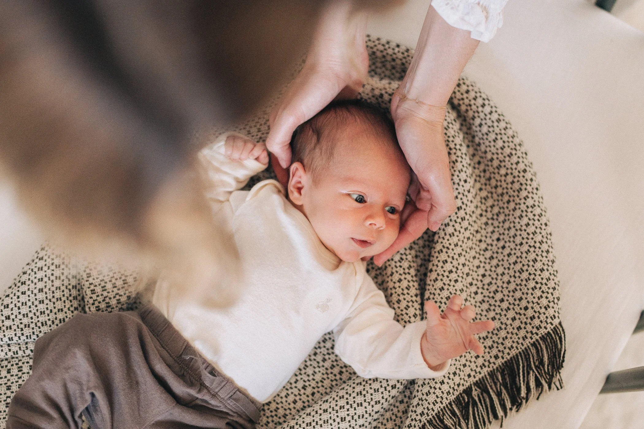 A newborn baby lying on a patterned blanket with a person's hands gently holding the baby's head, looking up.