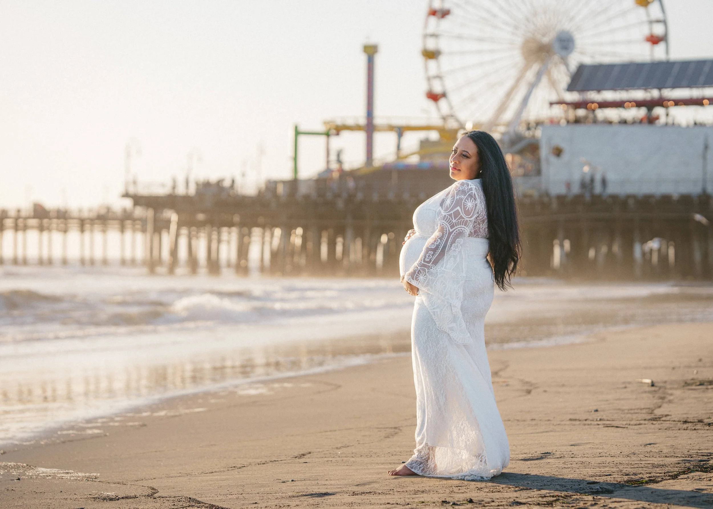 Pregnant woman in a white lace dress standing on the beach with a pier and Ferris wheel in the background at sunset.