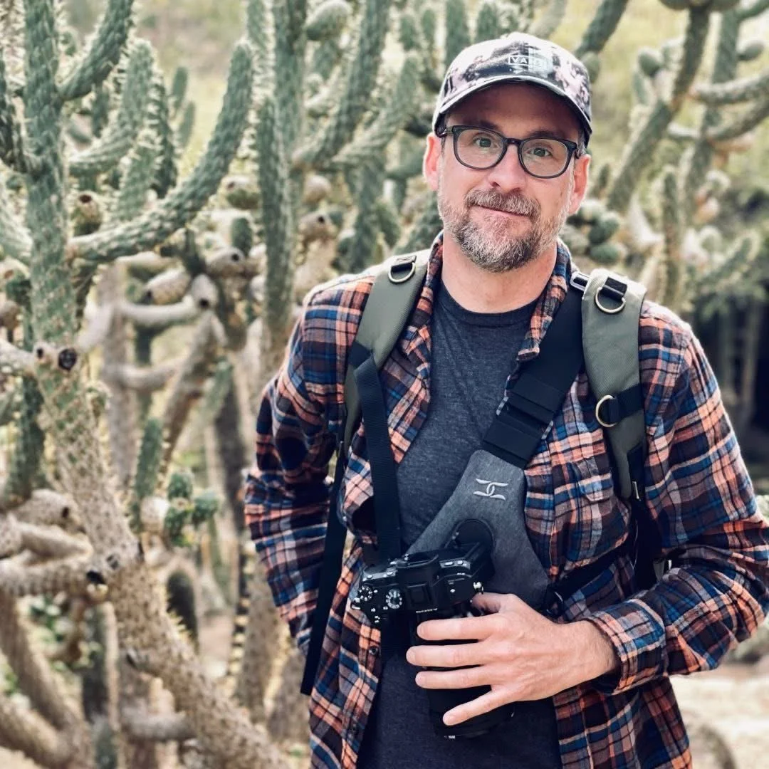 A man wearing glasses, a baseball cap, and a flannel shirt stands outdoors in front of cactus plants, holding a camera.