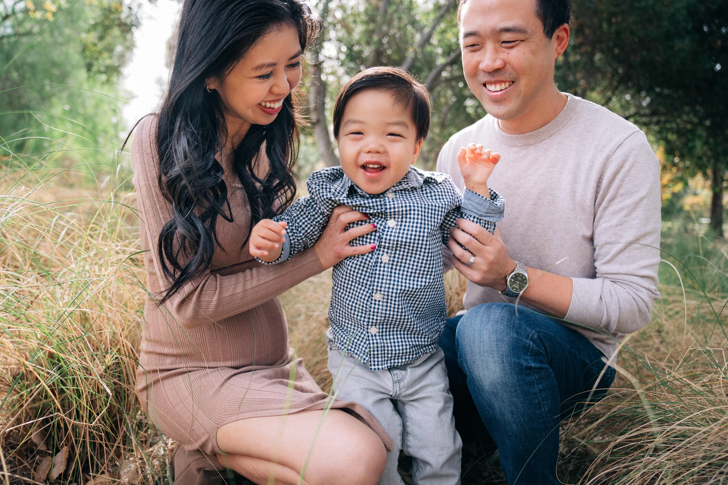 Happy Asian family of three outdoors in a grassy area, with a young boy smiling and standing between his smiling parents holding him by the hands.
