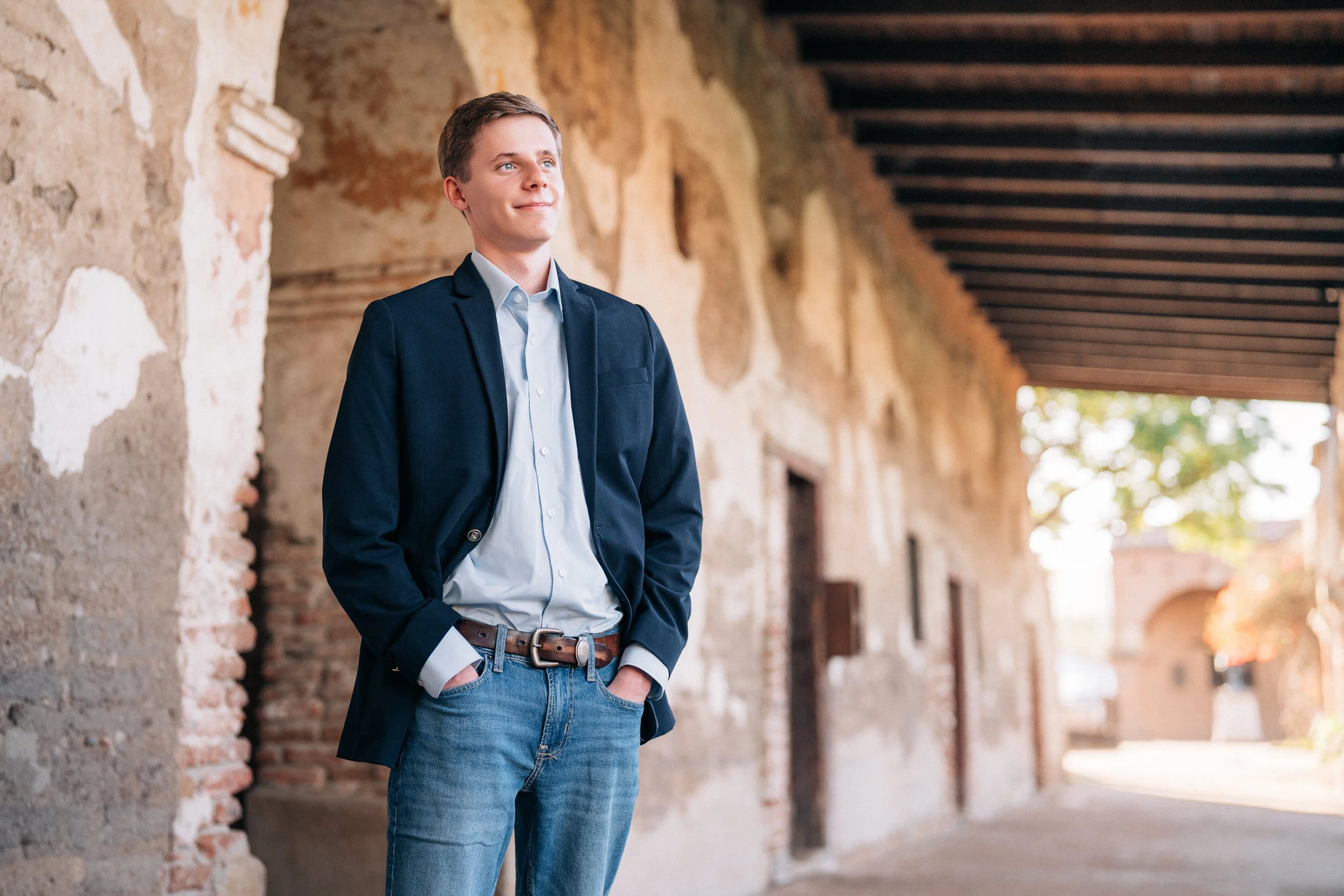 A young man in a navy blazer, light blue shirt, and jeans stands outdoors against an old, textured brick and stucco wall with a wooden overhang. He has a slight smile and hands in his pockets, looking upward.
