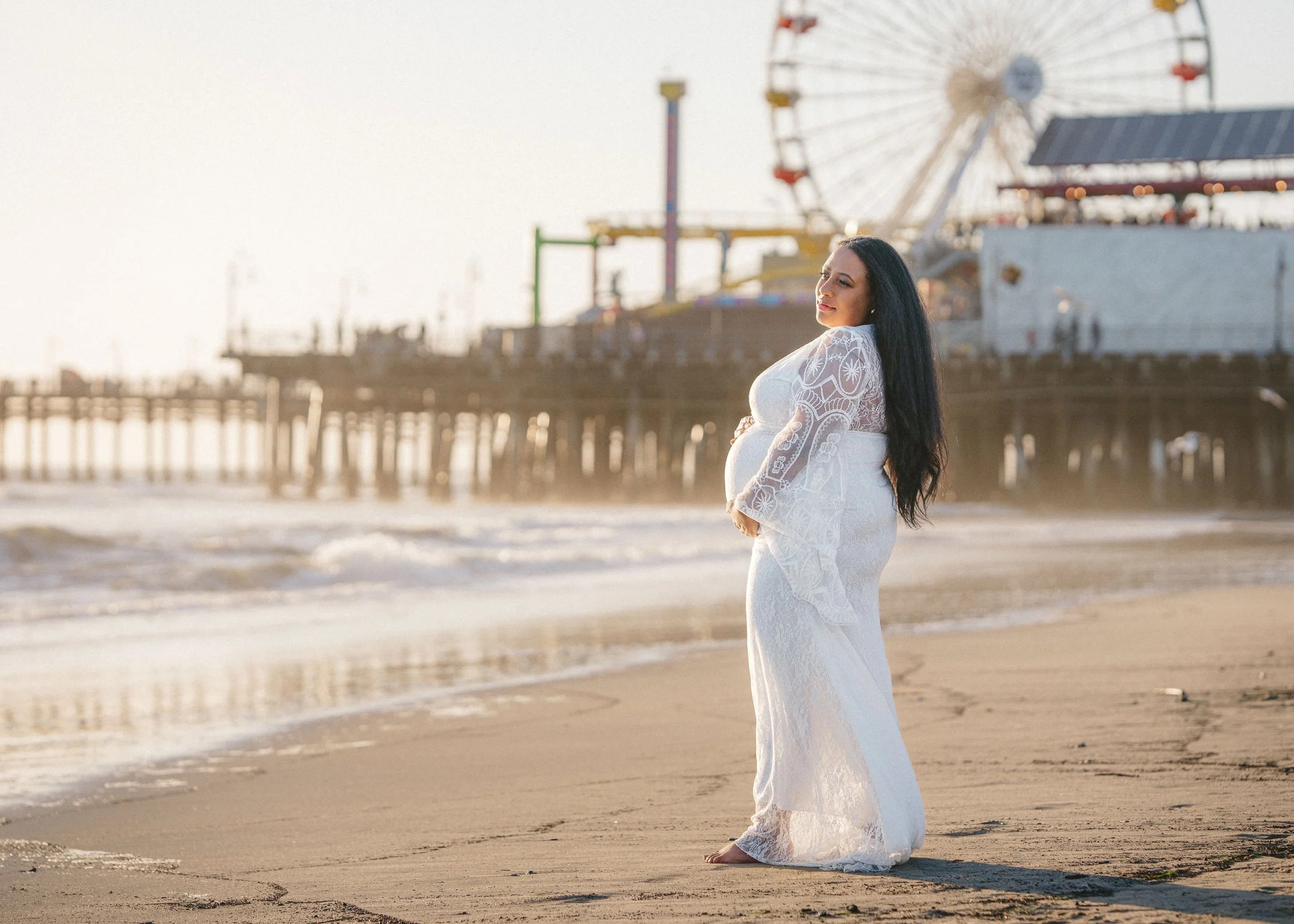 A pregnant woman in a white lace dress standing barefoot on the beach with a pier and amusement park rides in the background.