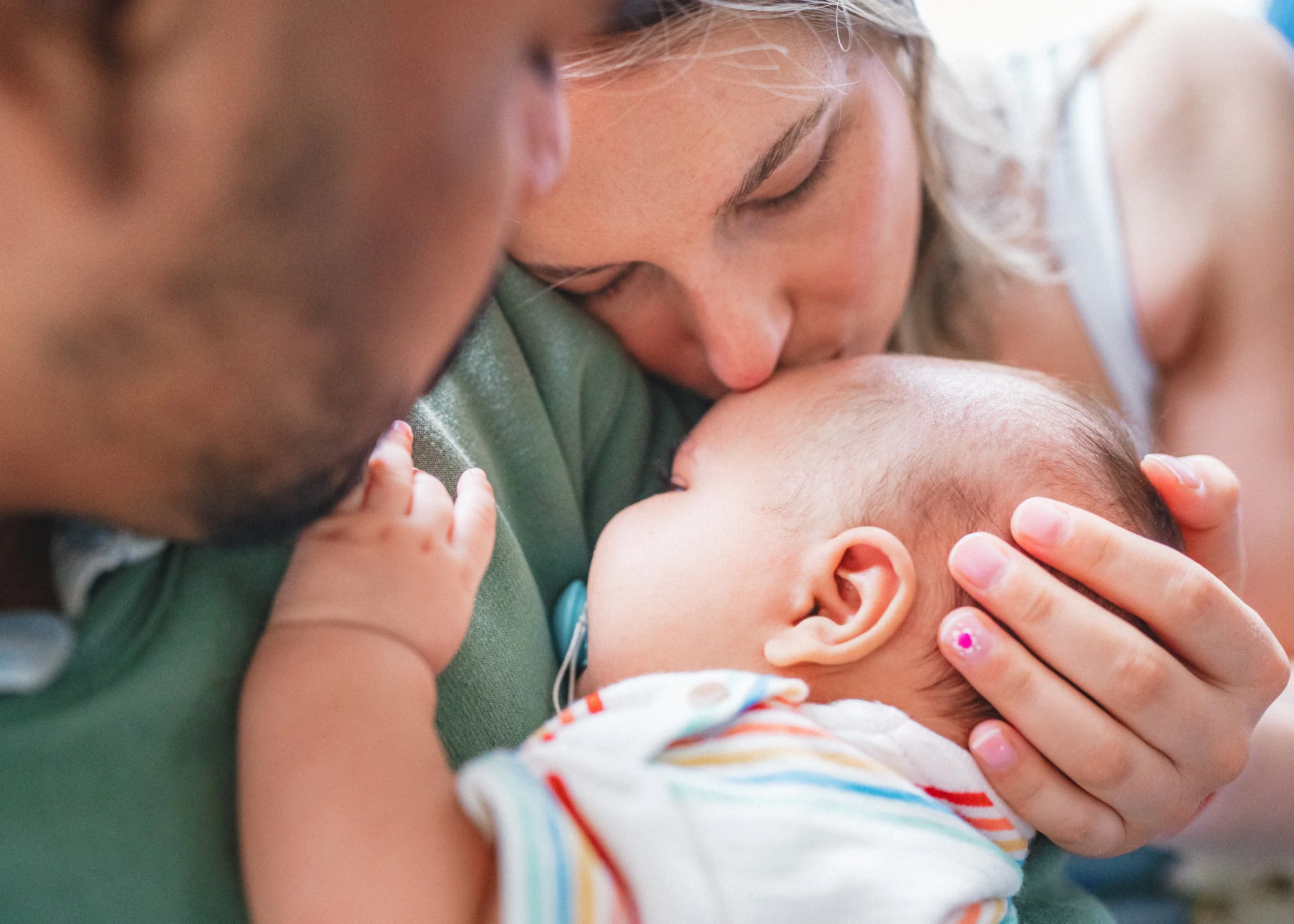 A woman holding a baby, kissing the baby's forehead, with a man close by, in a tender moment.